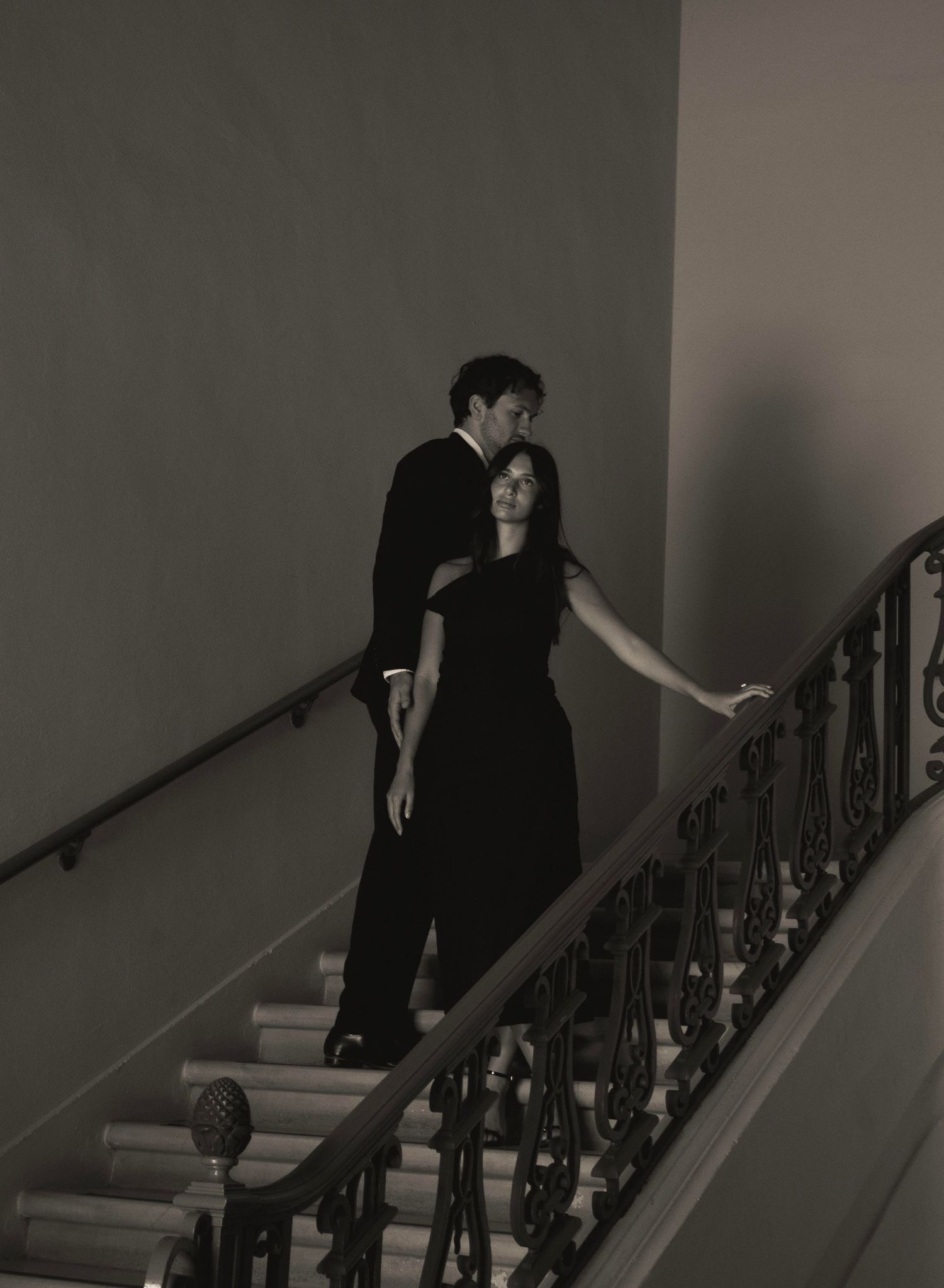 Elegant couple in black formal attire posing on ornate staircase in moody black and white photography.