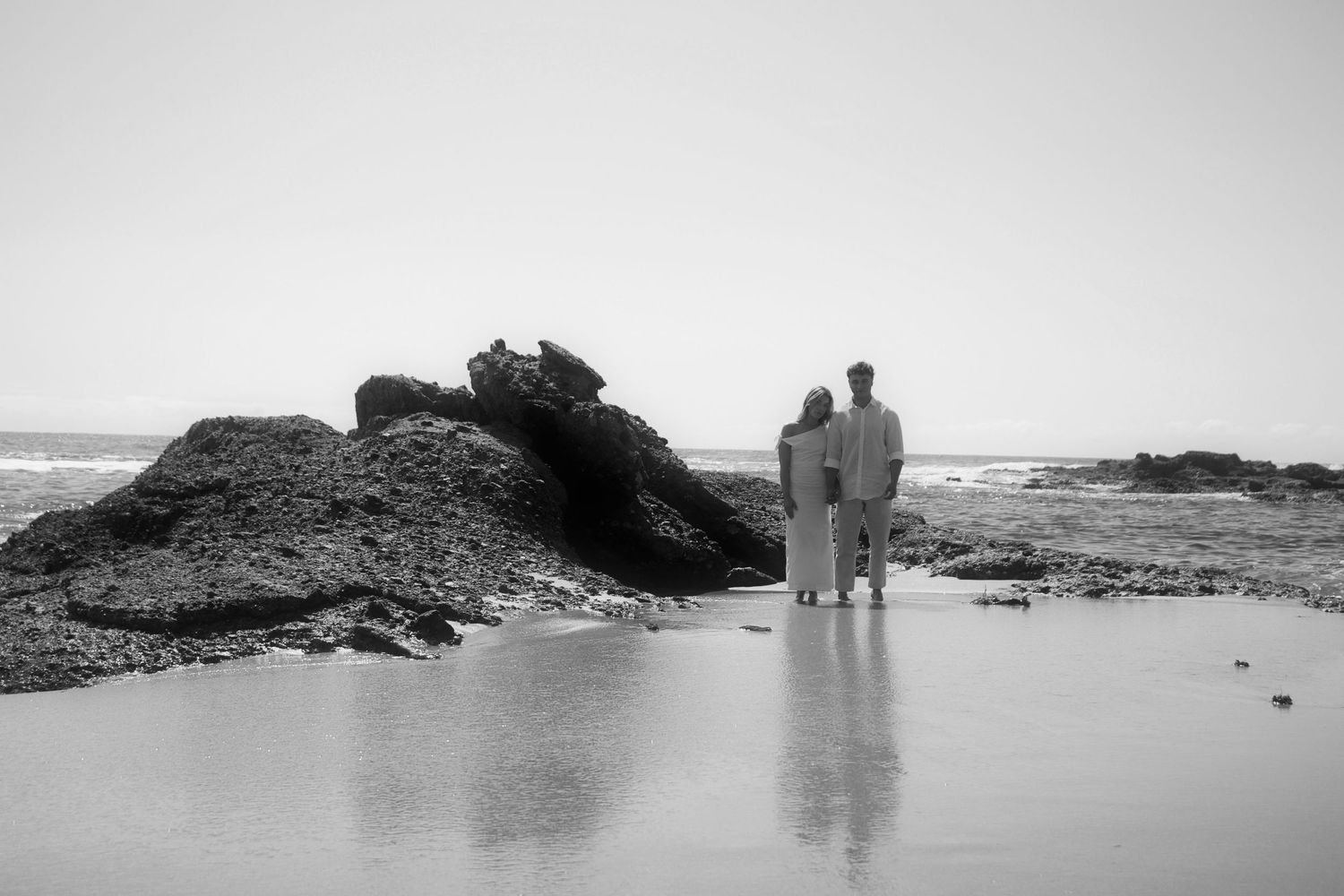 Black and white photo of couple standing together by ocean rocks on sandy beach.