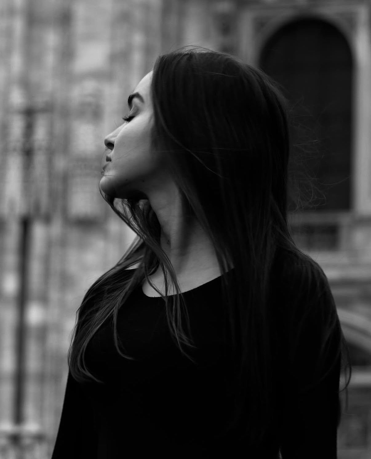 Black and white portrait of a young woman with long dark hair gazing upward outdoors near historic architecture.