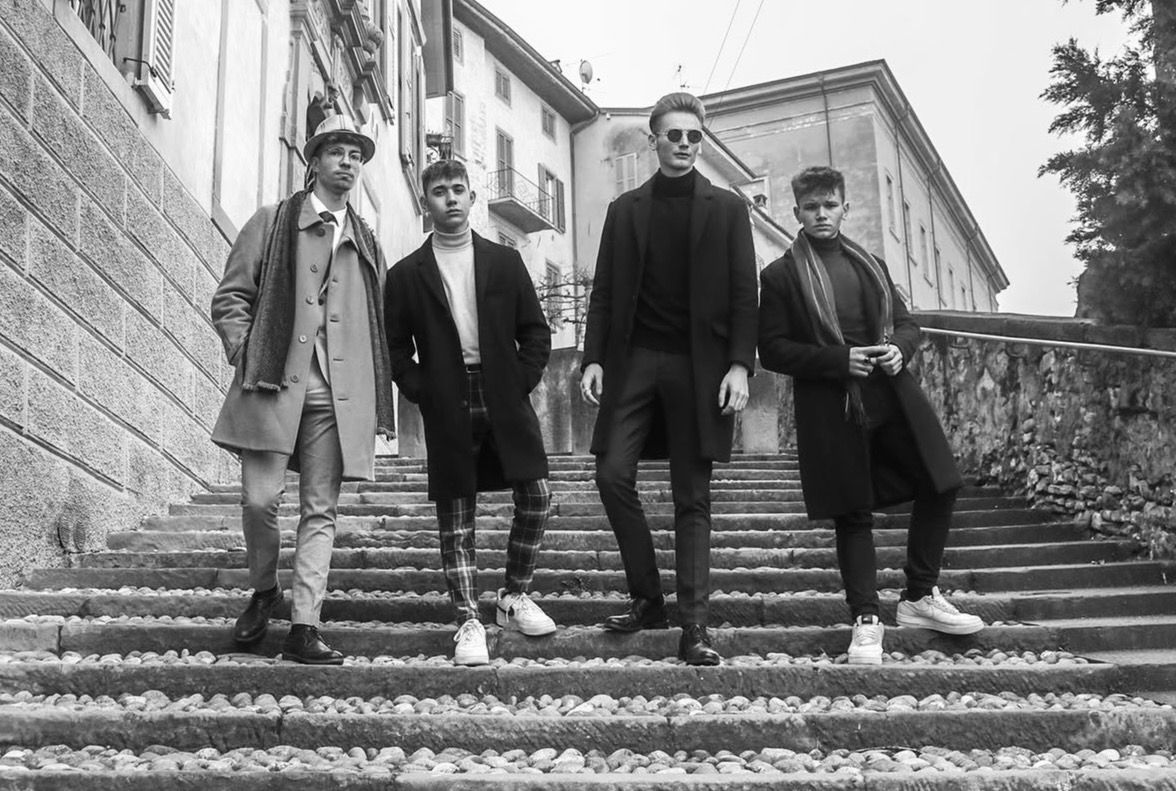 Four stylish men in black outfits pose on stone steps in a black and white urban photo.