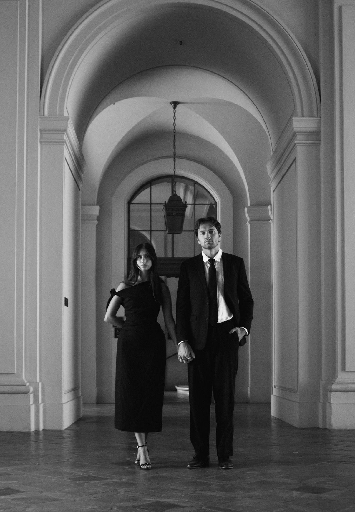 Black and white photo of an elegant couple in formal attire standing in a grand arched hallway.