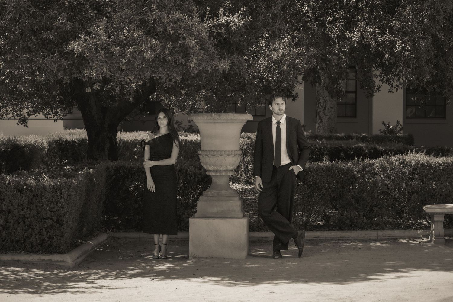 A man and woman in formal black attire pose separately in a sepia-toned garden with lush hedges and a stone urn.