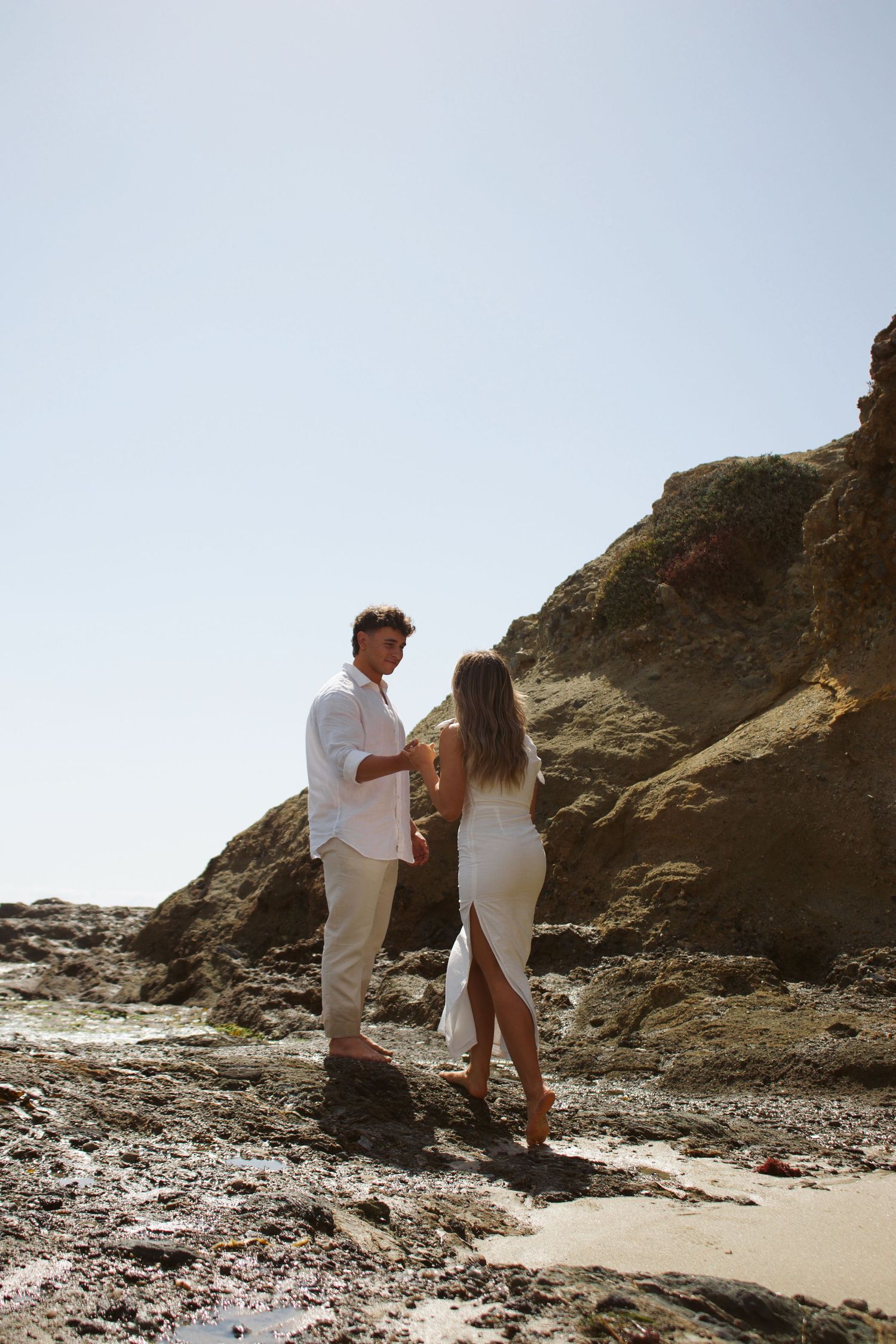 Couple holding hands near rugged rocky cliffs on a sunny beach with clear blue sky.