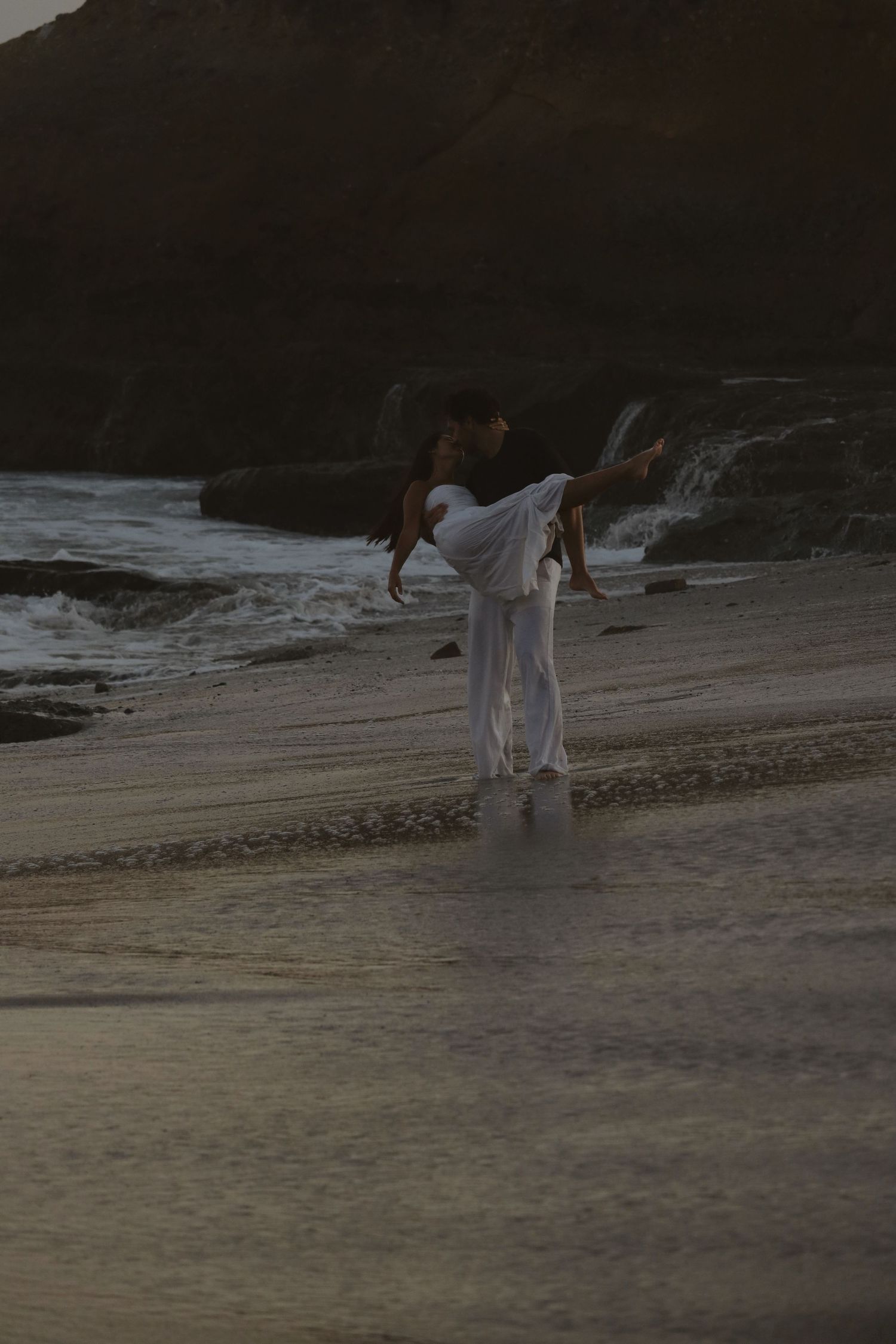 Couple in white attire dancing romantically on a misty beach at dusk with rocky cliffs in the background.