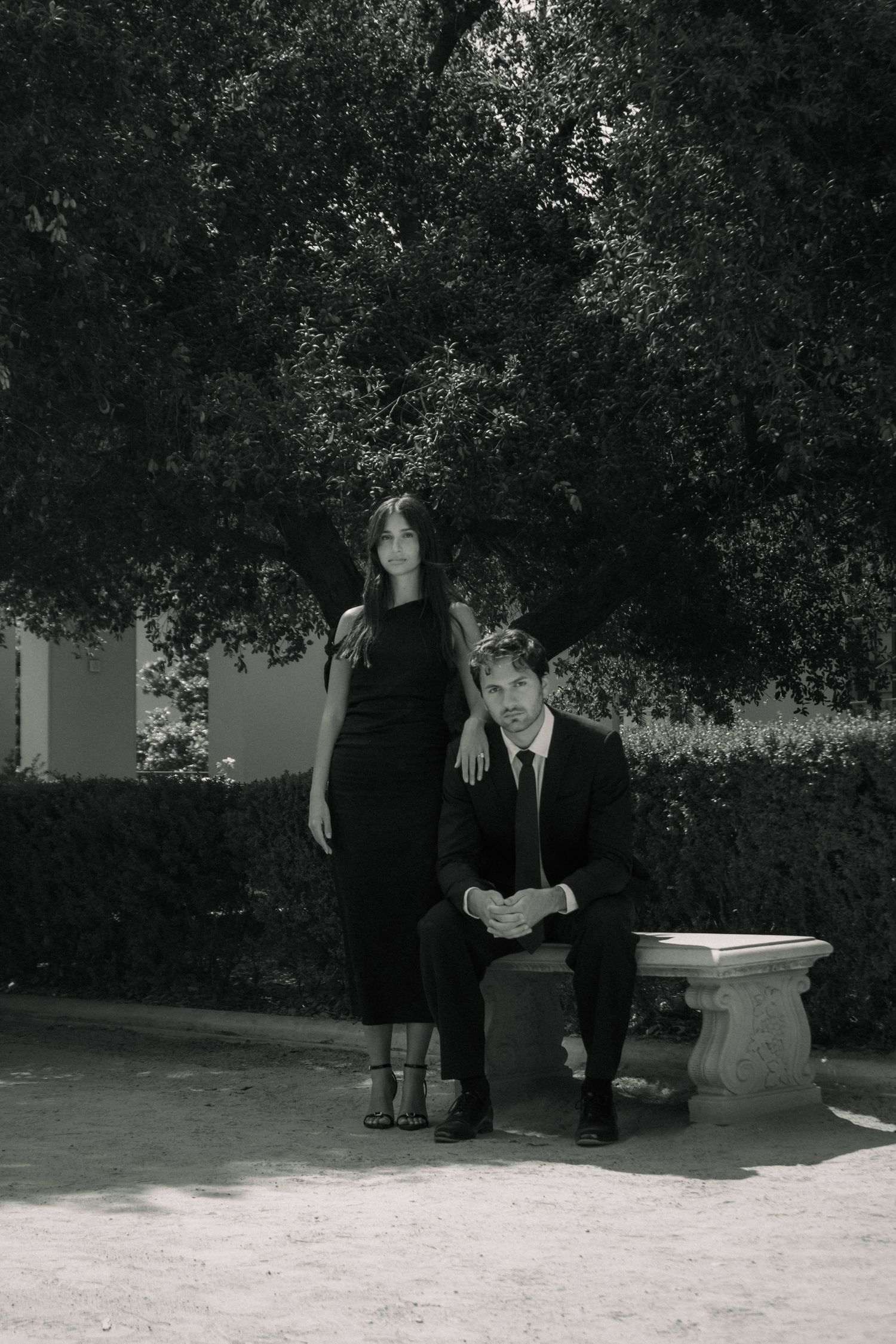 Black and white photo of a woman in black dress standing beside a suited man seated on a stone bench outdoors.