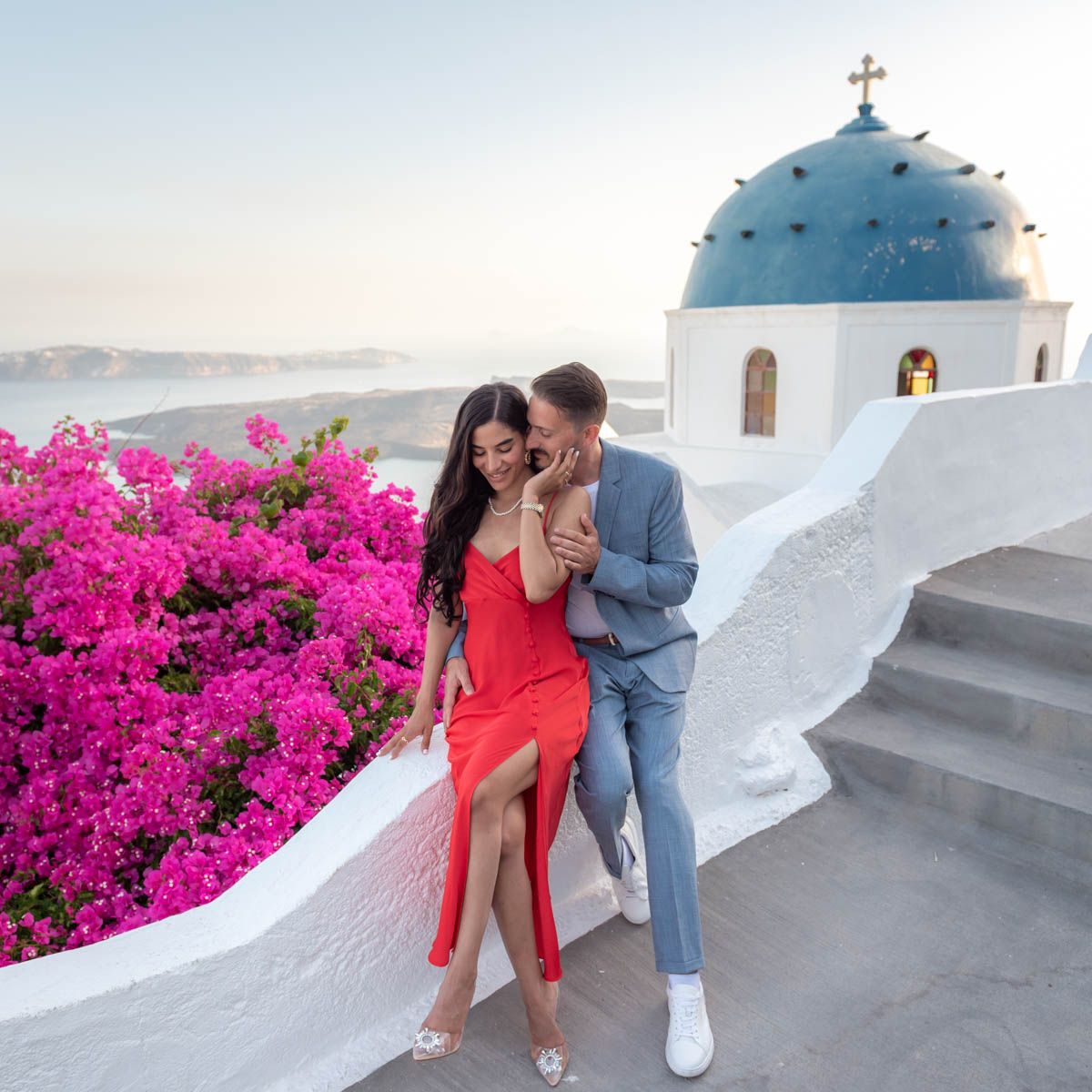 A couple sitting in Imerovigli village in Santorini during a photo shoot. Bougainvillea flowers and blue dome church in the background