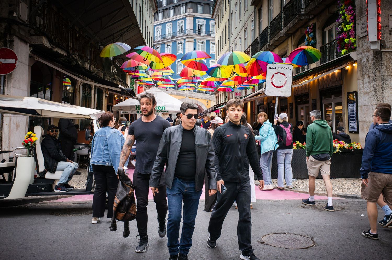 Pedestrians walk beneath colorful umbrellas on Lisbon's Pink Street, Portugal, in a vibrant historic street photography scene.