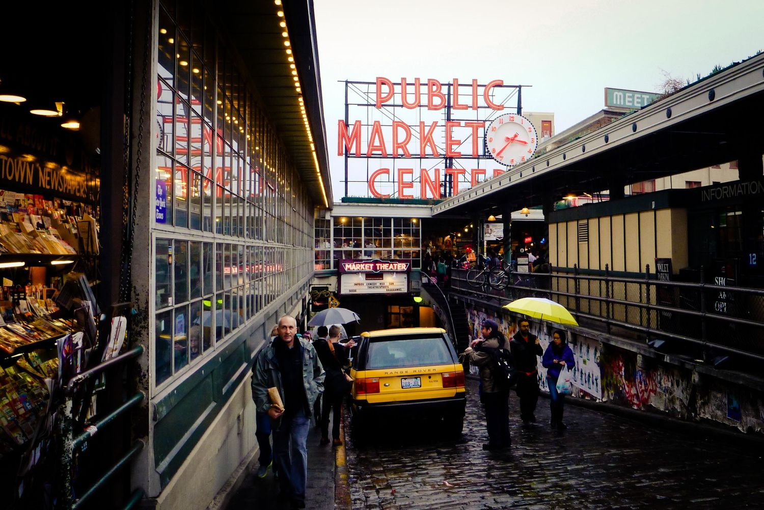 Rainy day at Seattle's Pike Place Market with the Public Market Center sign, umbrellas, and pedestrians on cobblestone streets.