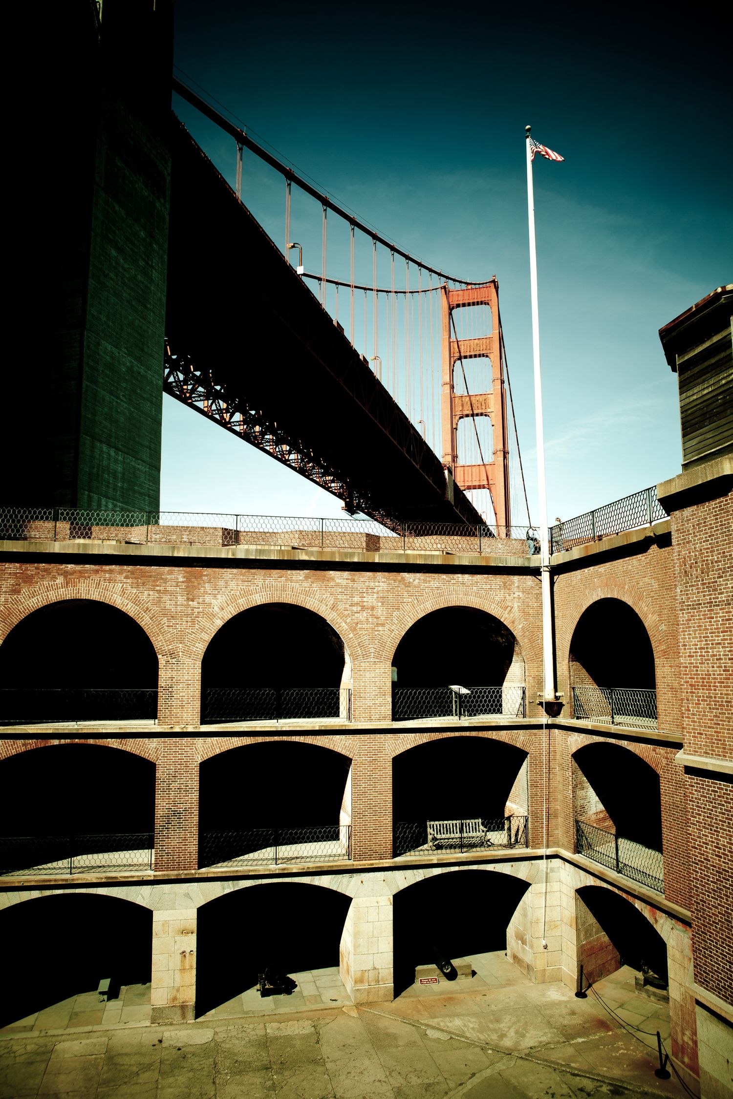 Golden Gate Bridge towering above Fort Point's brick arches and American flag in San Francisco, viewed from below.