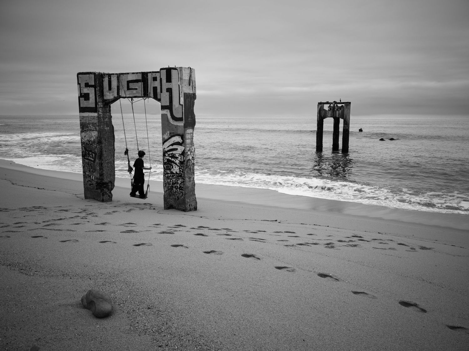 Person on swing beneath Davenport Beach ruins in California, with ocean waves and coastal remains in dramatic black and white.