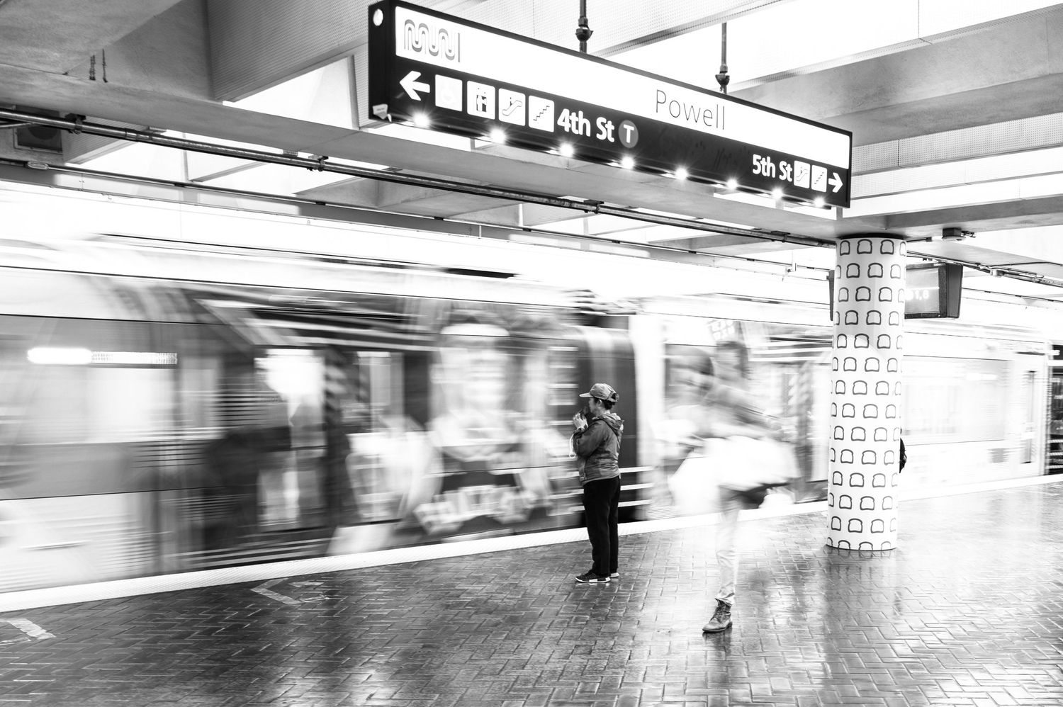 Motion-blurred BART train passes a waiting commuter at San Francisco's Powell Station in a black-and-white street photograph.