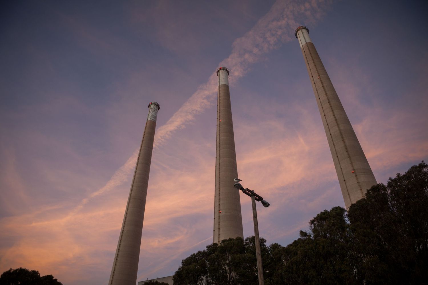 Three iconic Morro Bay power plant chimney stacks rise into a colorful California sunset with dramatic clouds and coastal silhouettes.