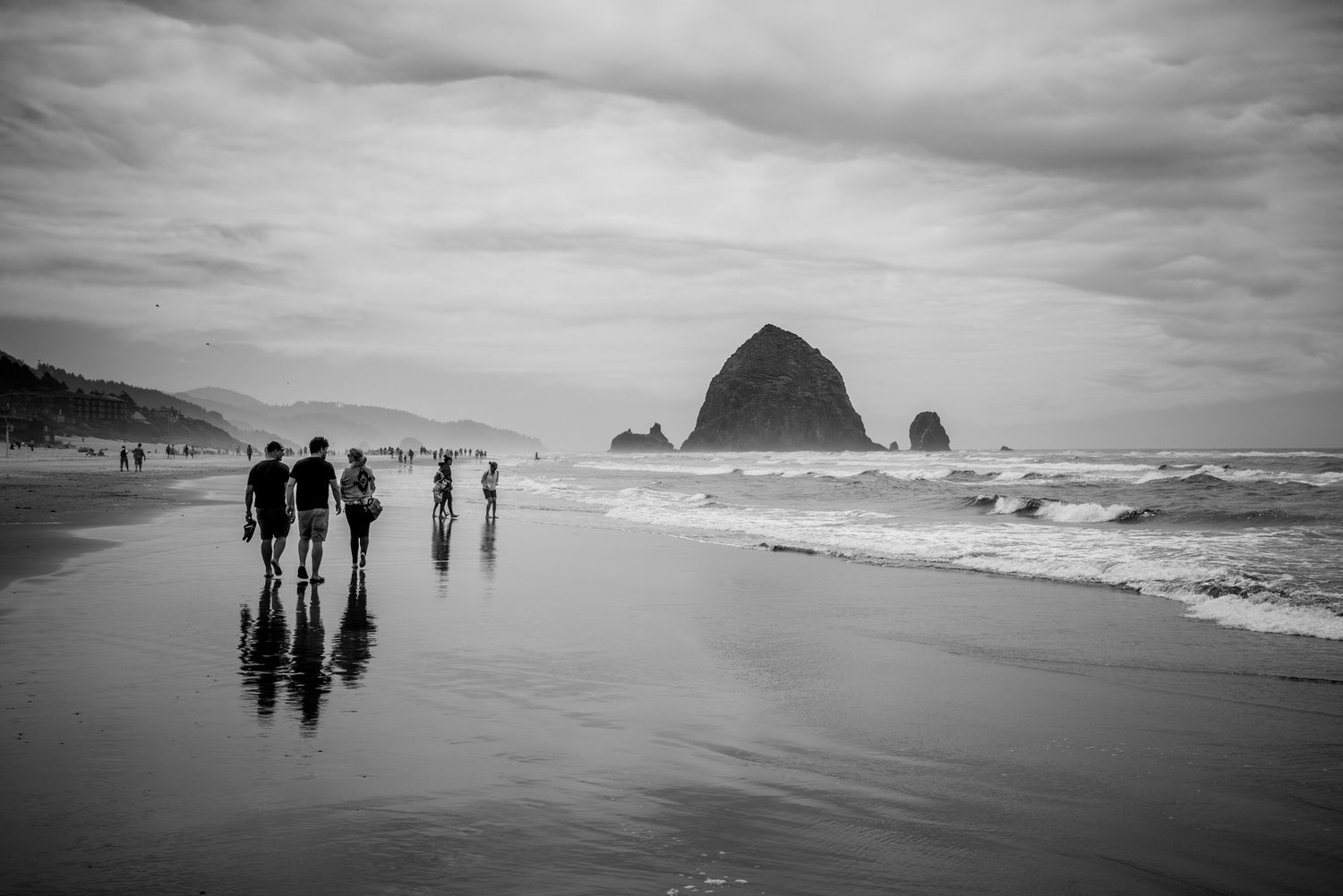 Black and white view of Haystack Rock at Cannon Beach, Oregon, with walkers, reflections, waves, and dramatic coastal clouds.