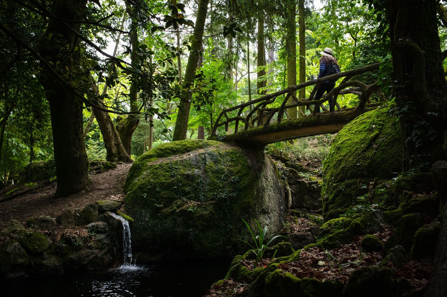 Woman in a white hat crossing a rustic bridge in Quinta da Regaleira, Sintra, Portugal, surrounded by lush moss and forest.