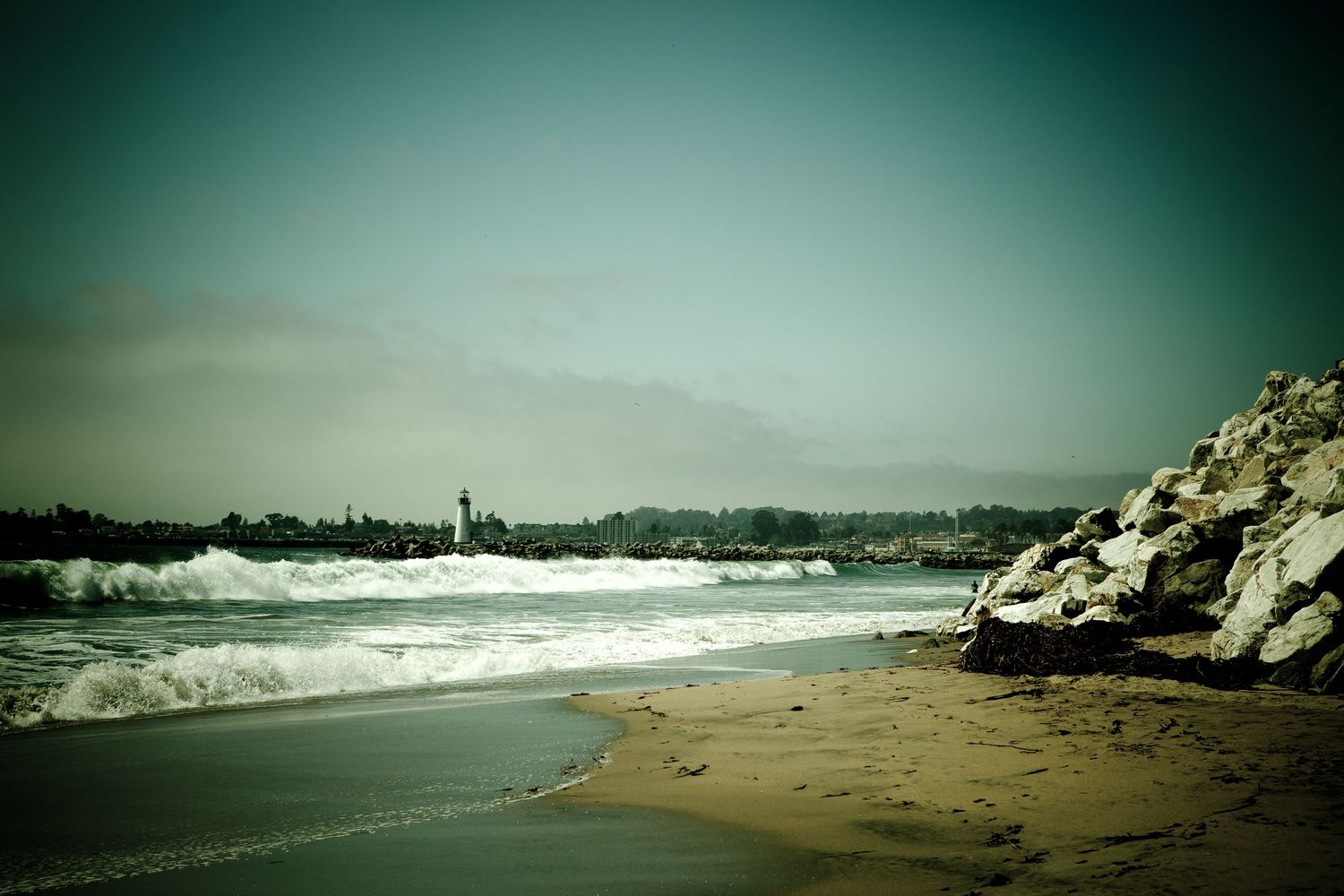 Breaking waves along Santa Cruz Beach with the Santa Cruz Harbor Lighthouse and rocky jetty on the California coast.