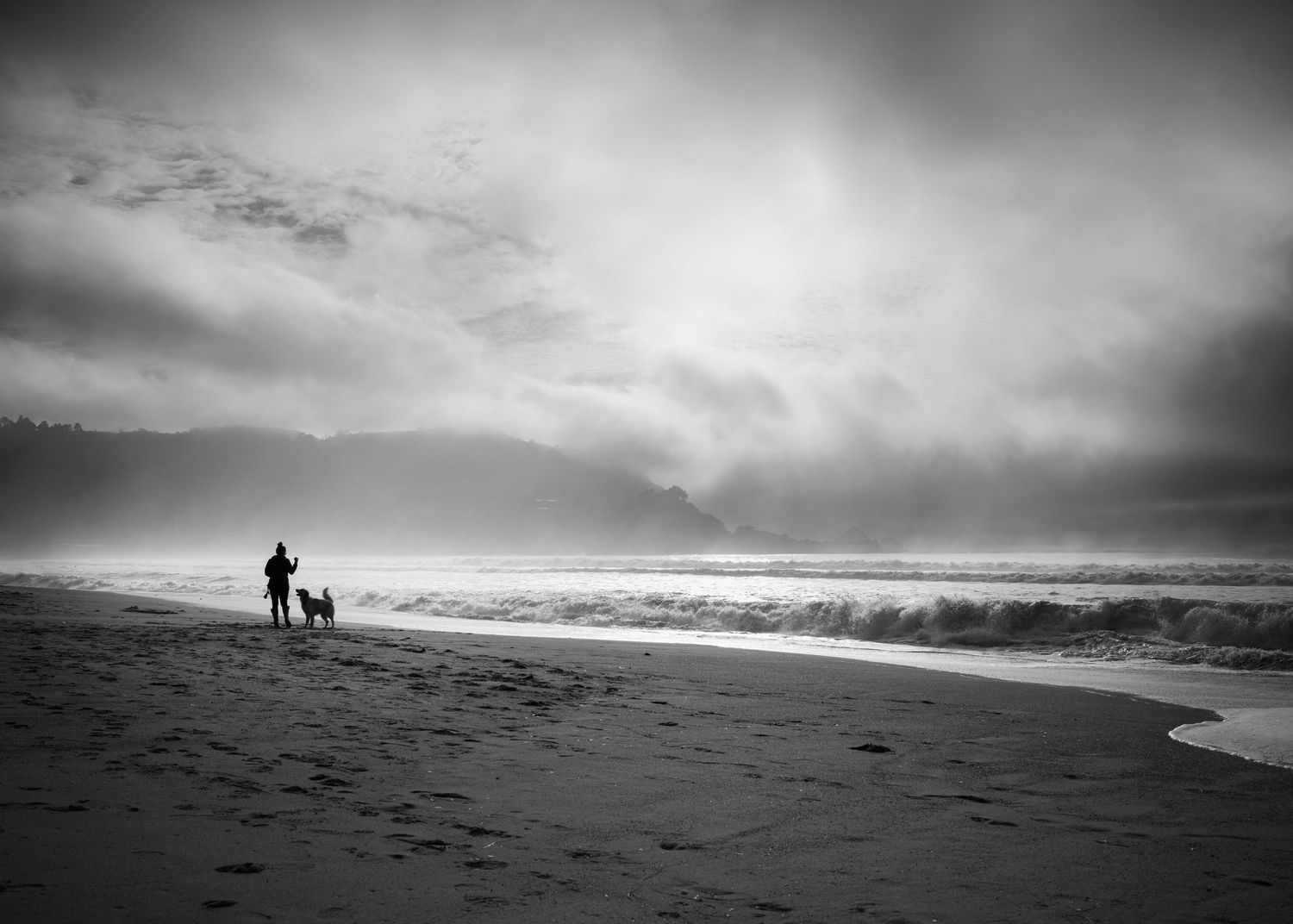 Person walking a dog beneath dramatic storm light at Pacifica State Beach, California, in a moody black and white coastal scene.