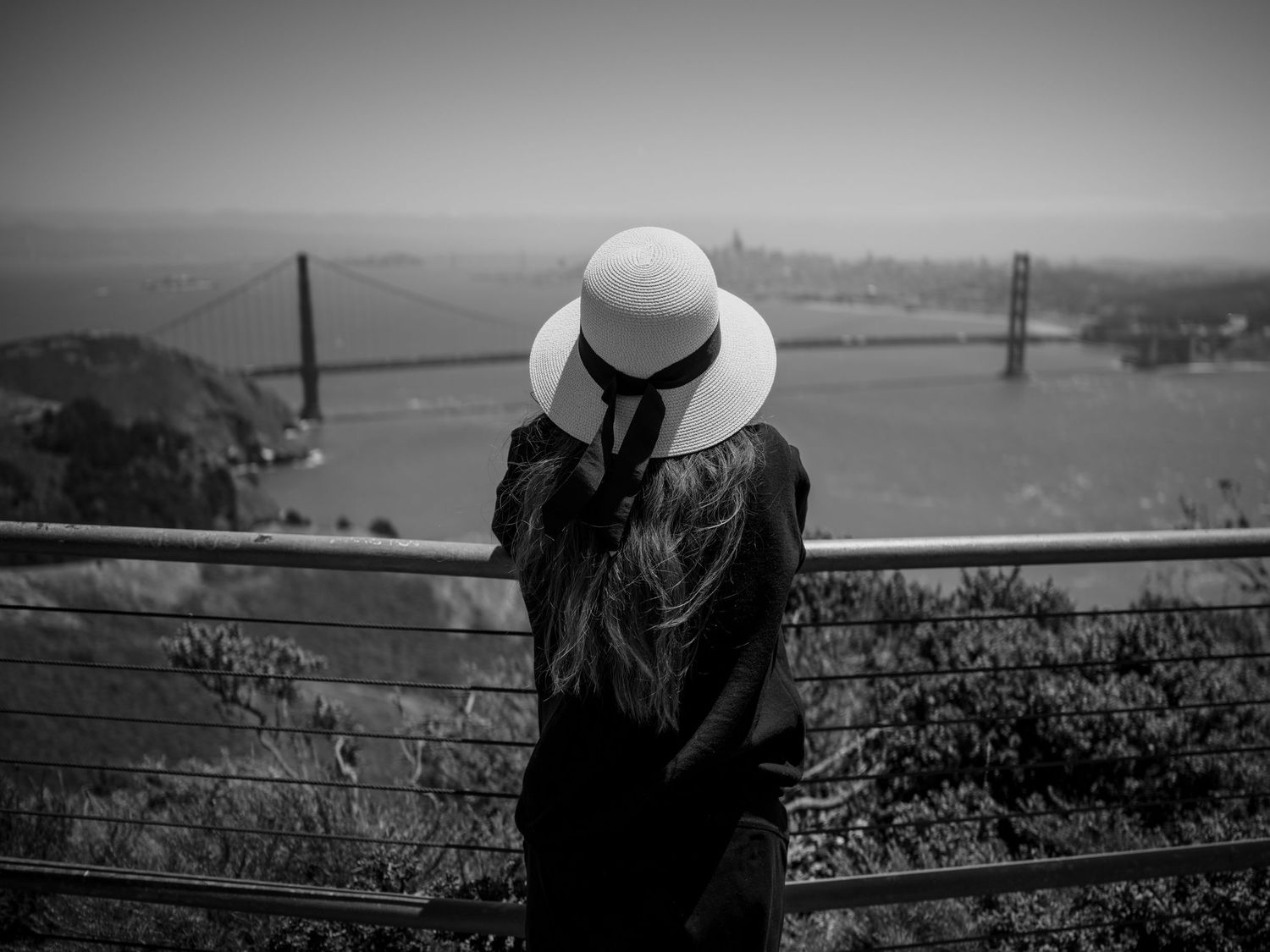 Woman in a white sun hat overlooks the Golden Gate Bridge from Hawk Hill, Marin Headlands, San Francisco in black and white.