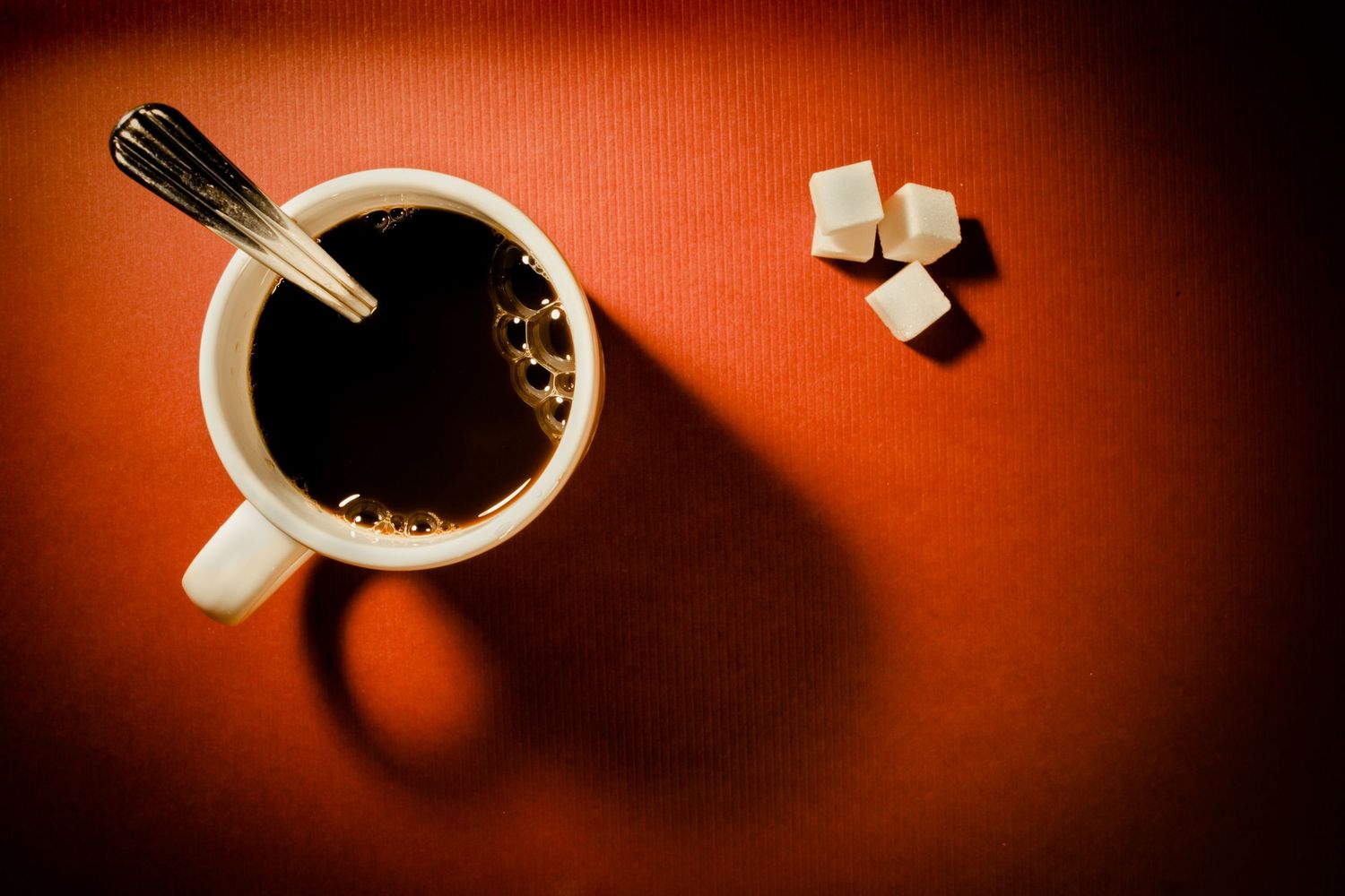 Minimalist fine art still life of a black coffee cup casting long shadows beside sugar cubes on a warm red background.