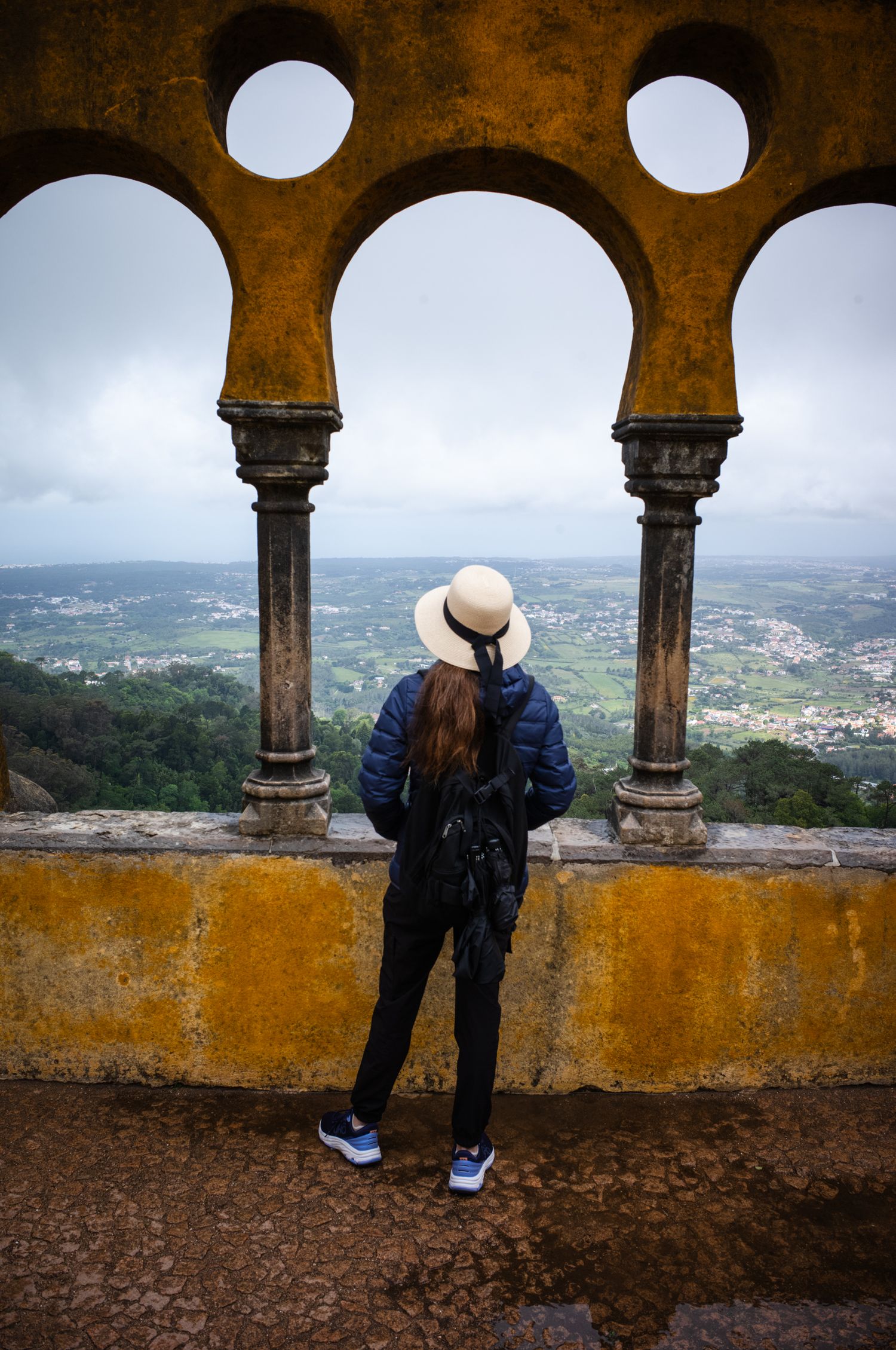 Woman in a white hat framed by Pena Palace arches overlooking the Sintra landscape, Portugal, from the iconic palace viewpoint.