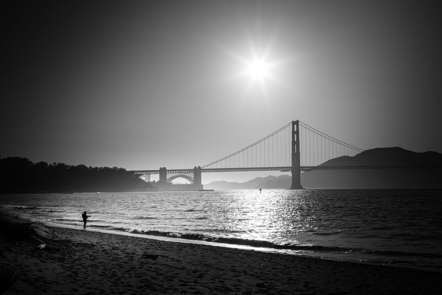 Black and white view of the Golden Gate Bridge with a starburst sun, shimmering bay, and lone figure on the beach in San Francisco.