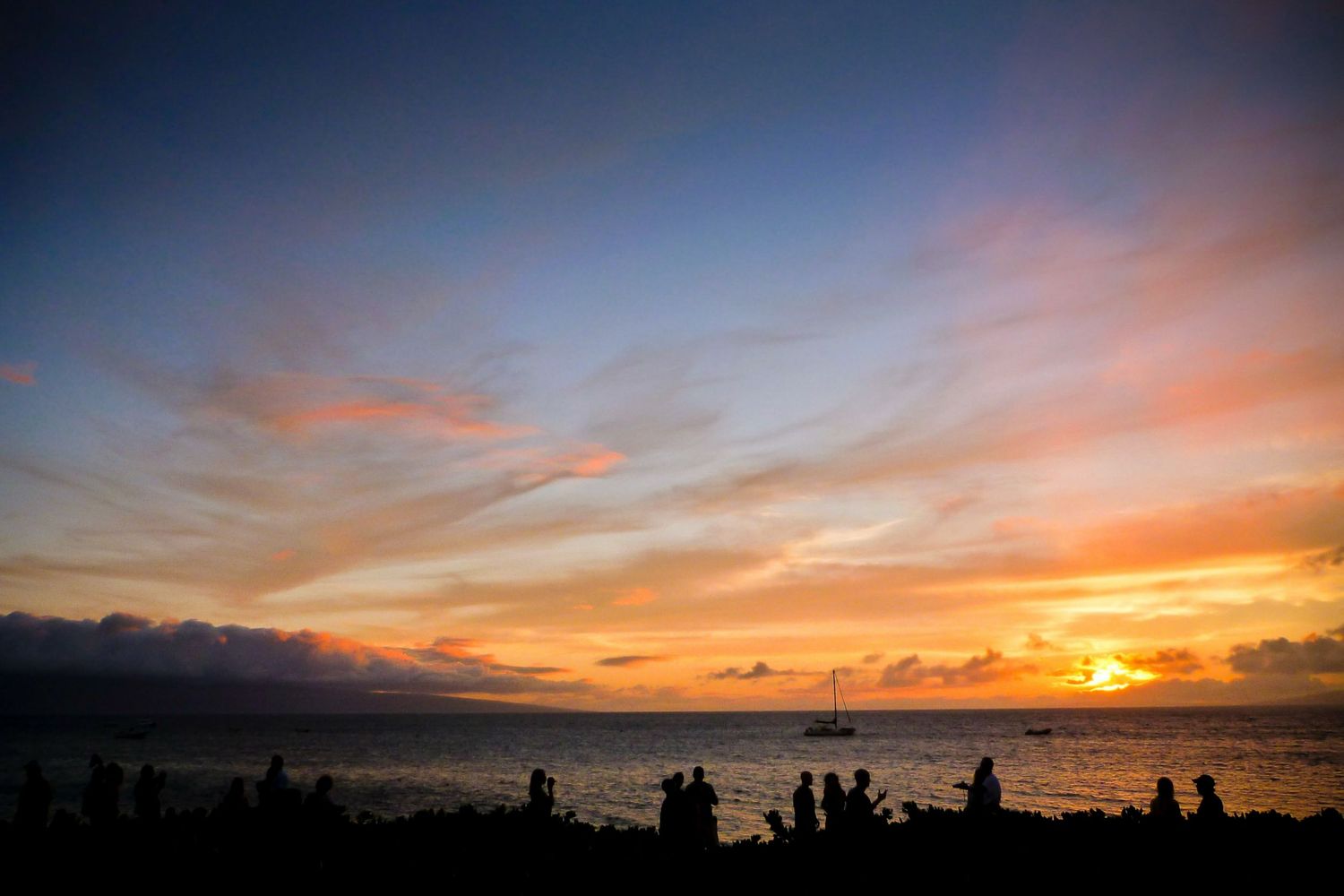 Silhouetted visitors watch a vibrant Maui sunset with a sailboat offshore beneath colorful tropical skies over the Pacific Ocean.