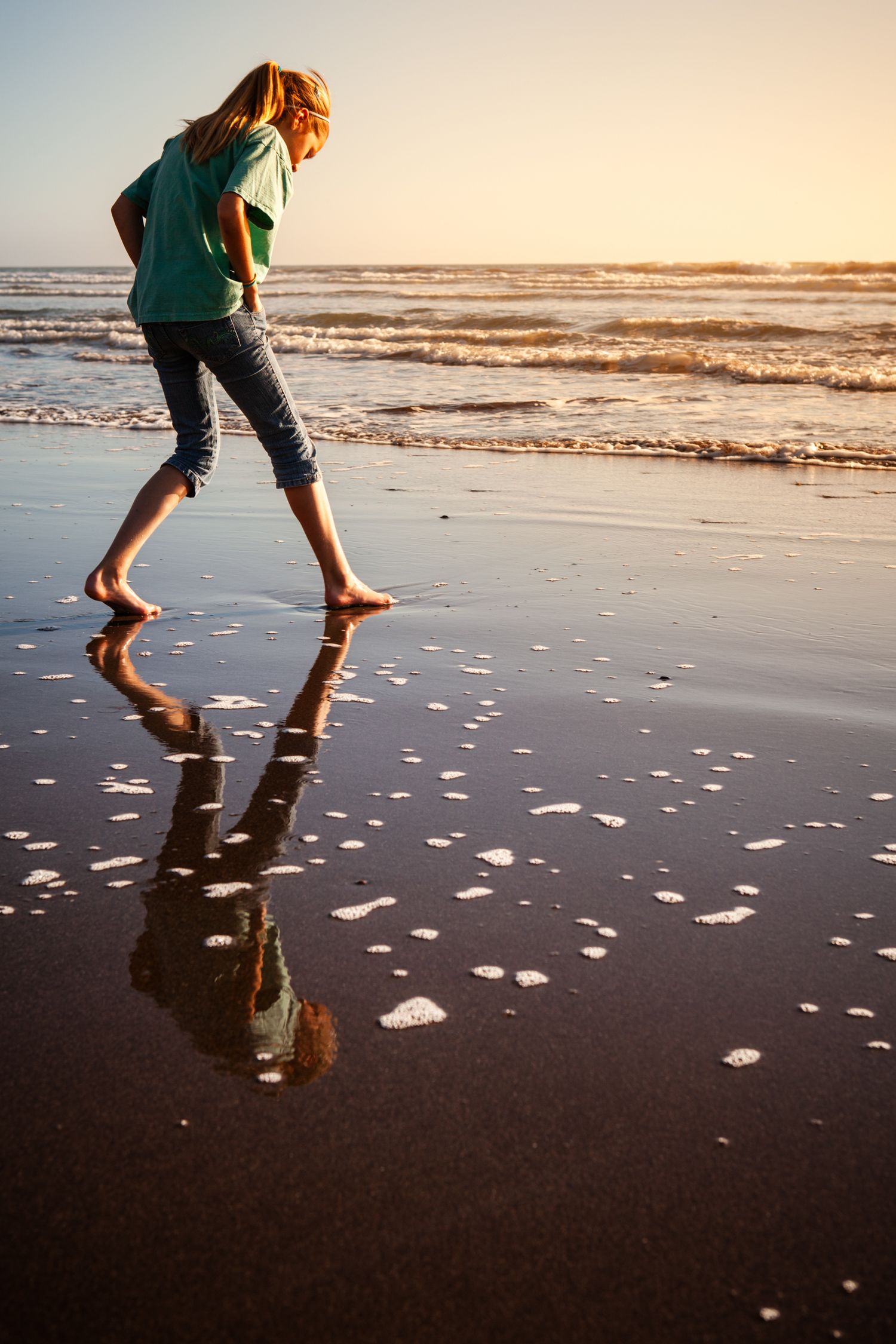 Barefoot child walking along the shoreline at sunset in Santa Cruz, California, reflected in wet sand beneath golden coastal light.