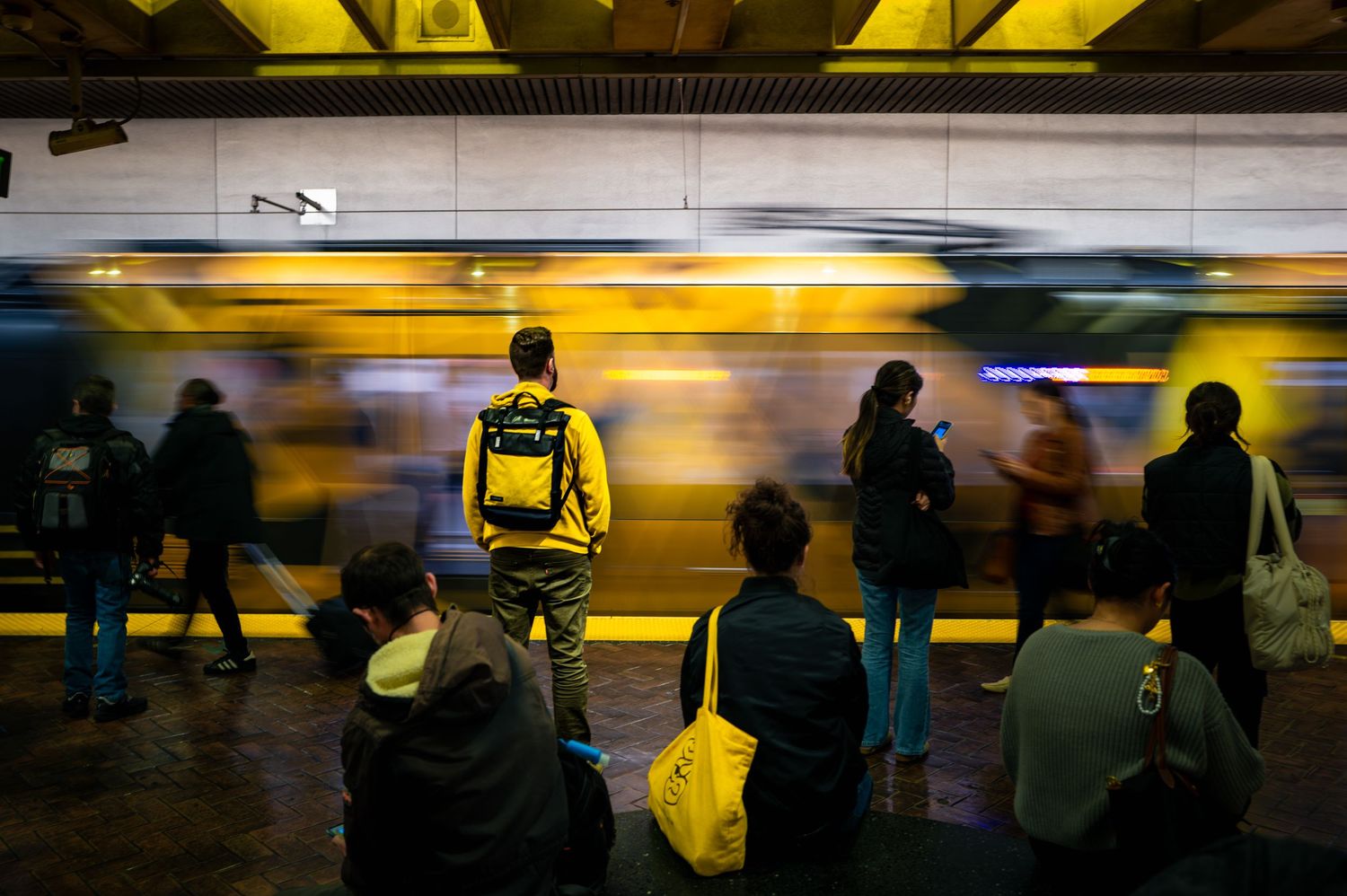 Commuter in yellow waits as a speeding BART train blurs through a San Francisco station, capturing the rhythm of urban life.