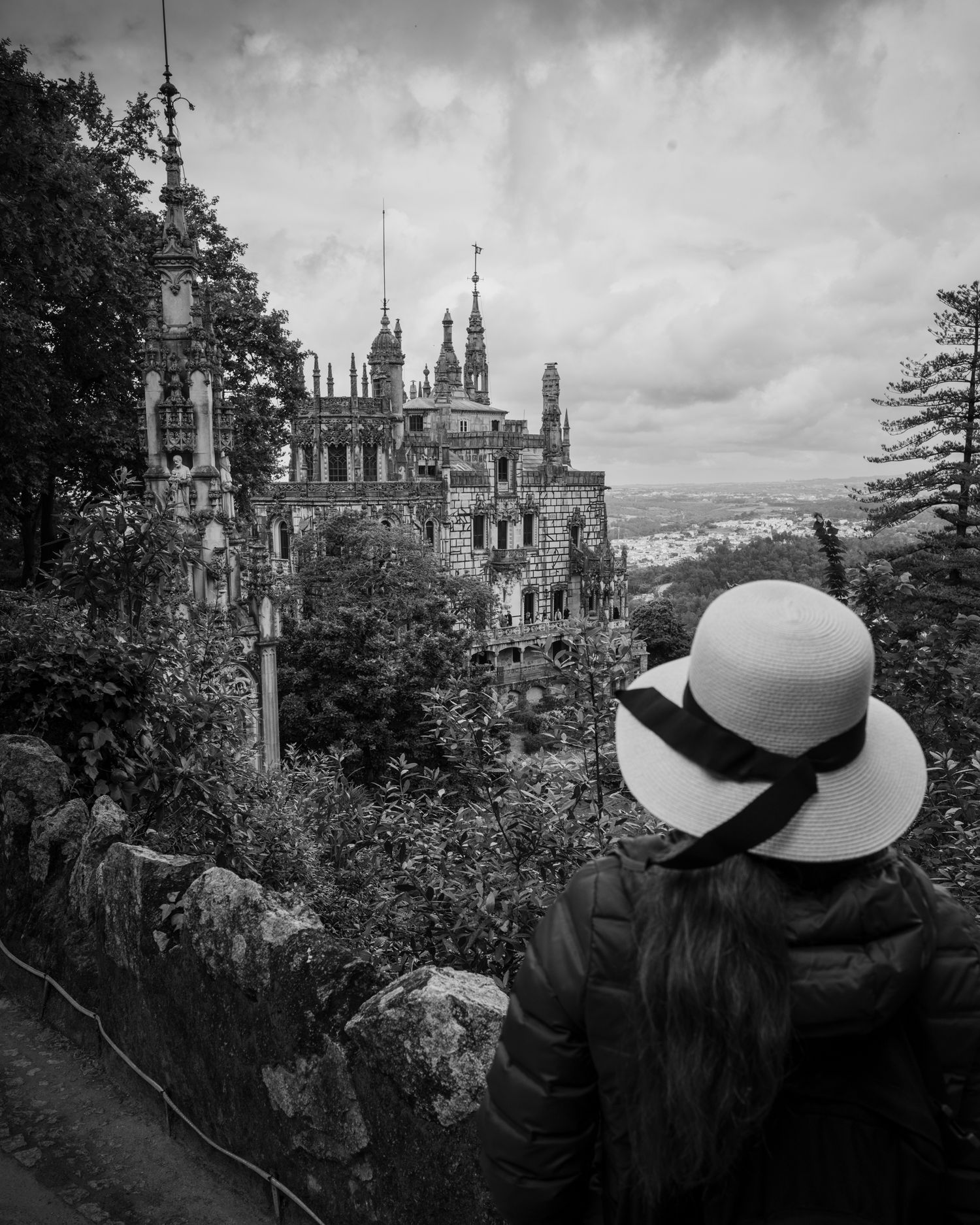 Woman in white hat overlooking Quinta da Regaleira in Sintra, Portugal beneath dramatic skies in a black and white travel photograph.
