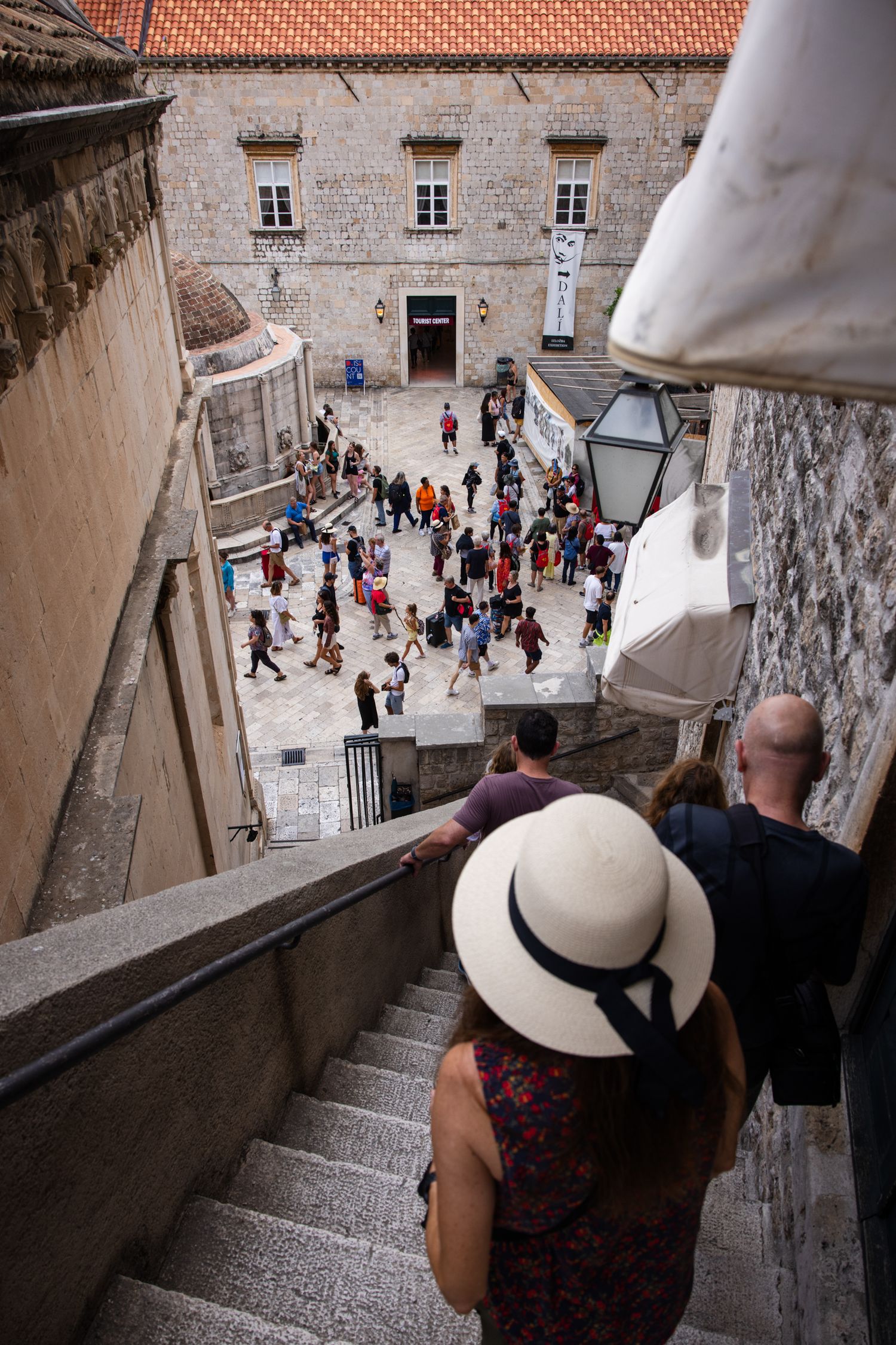 Woman in a white sun hat descends stone steps from Dubrovnik's Old Town walls overlooking the historic streets of Croatia.