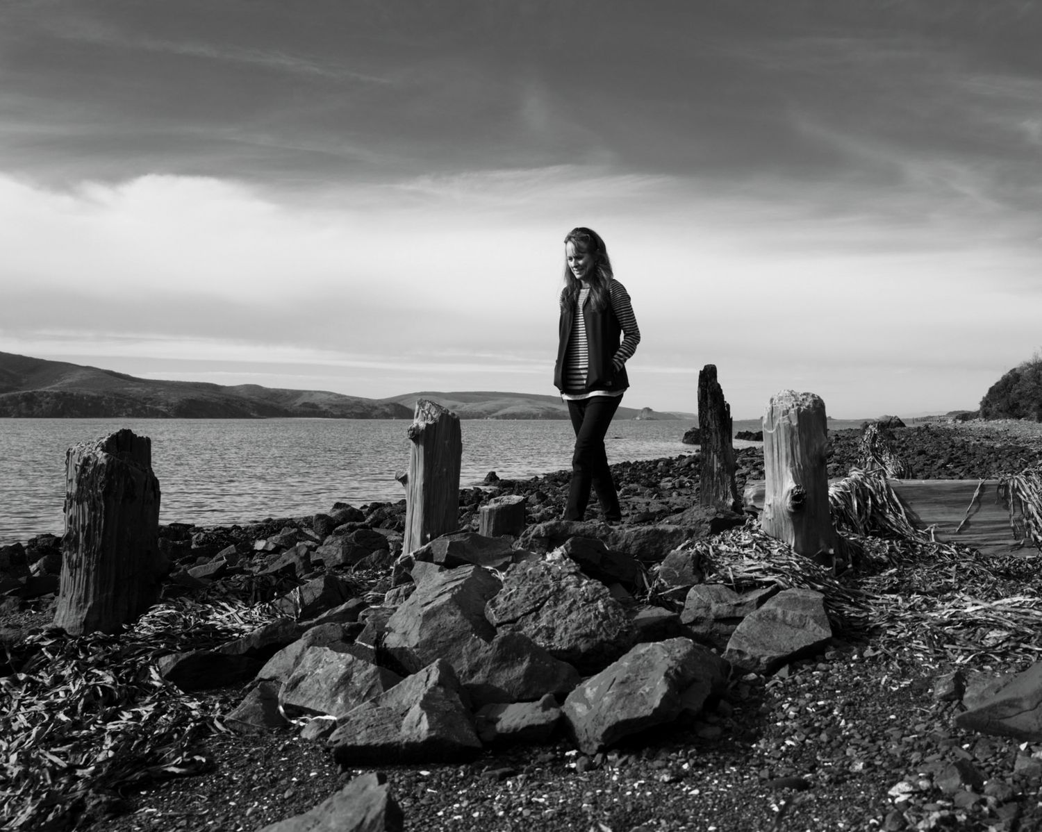 Woman walking among weathered pilings on the rocky shoreline of Tomales Bay, California beneath dramatic skies in black and white.