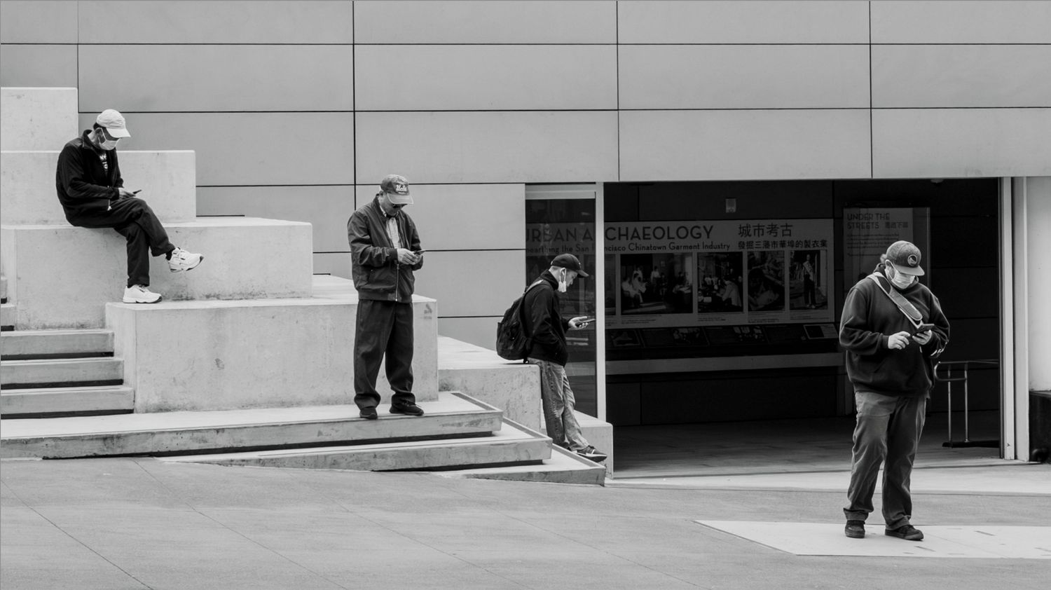 Four people absorbed in smartphones outside a modern building in San Francisco, captured in a minimalist black and white street photograph.
