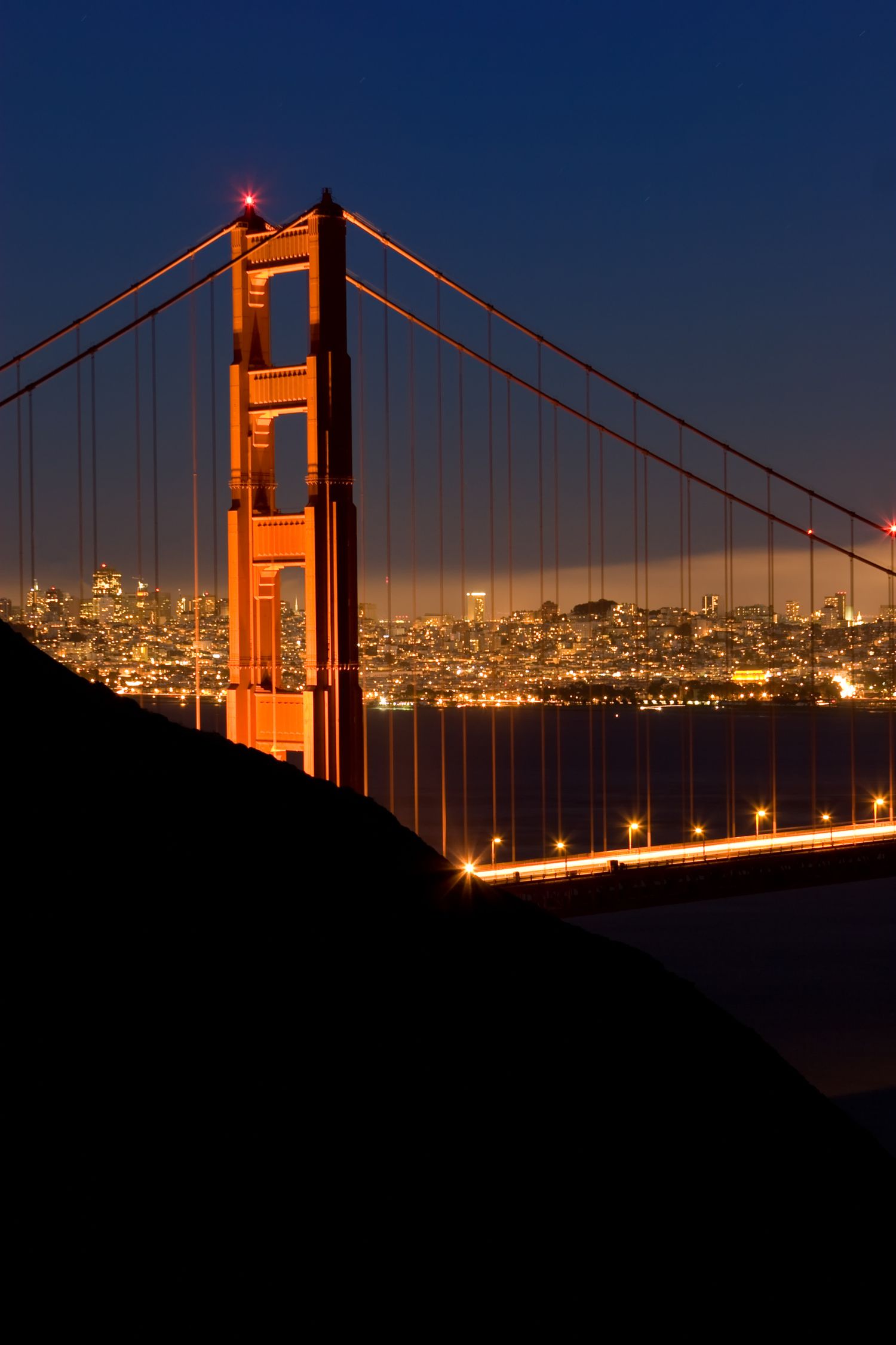 Illuminated Golden Gate Bridge tower at blue hour with the San Francisco skyline glowing across the bay in a dramatic night scene.