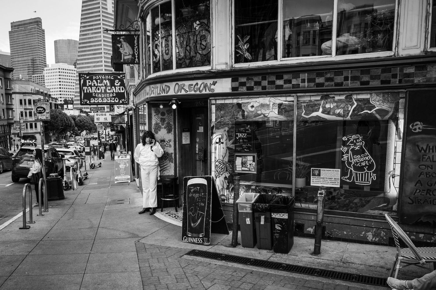 Black and white San Francisco street scene with Vesuvio Cafe, vintage storefronts, pedestrians, and downtown skyline.