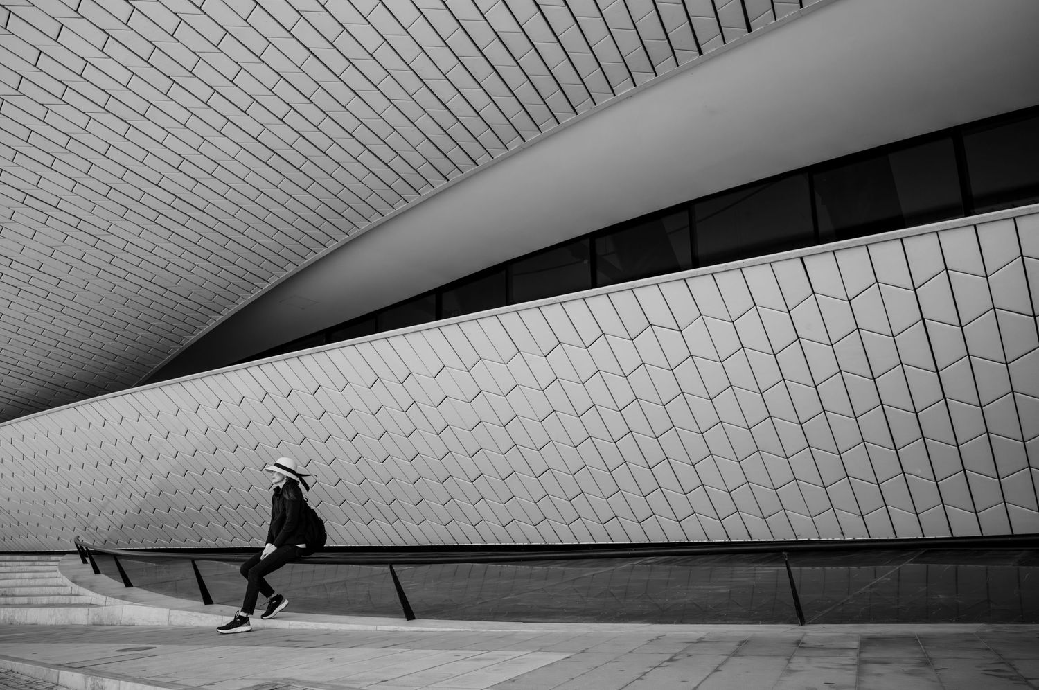 Woman in a white hat beneath the sweeping curves of Lisbon's MAAT Museum, a minimalist black-and-white image from The Hat Series.
