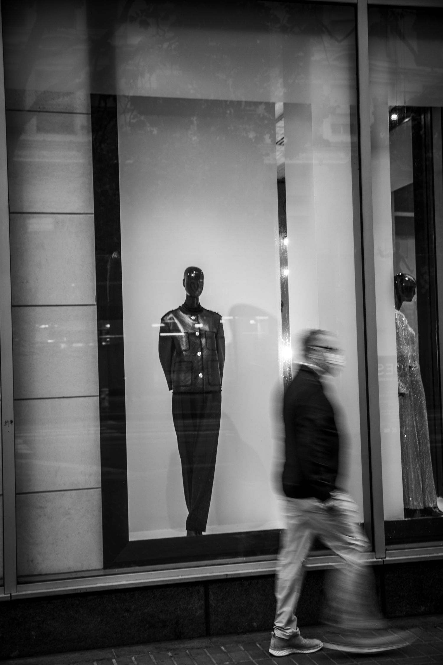 Motion-blurred man walks past a mannequin in a San Francisco storefront, capturing urban life in black-and-white street photography.