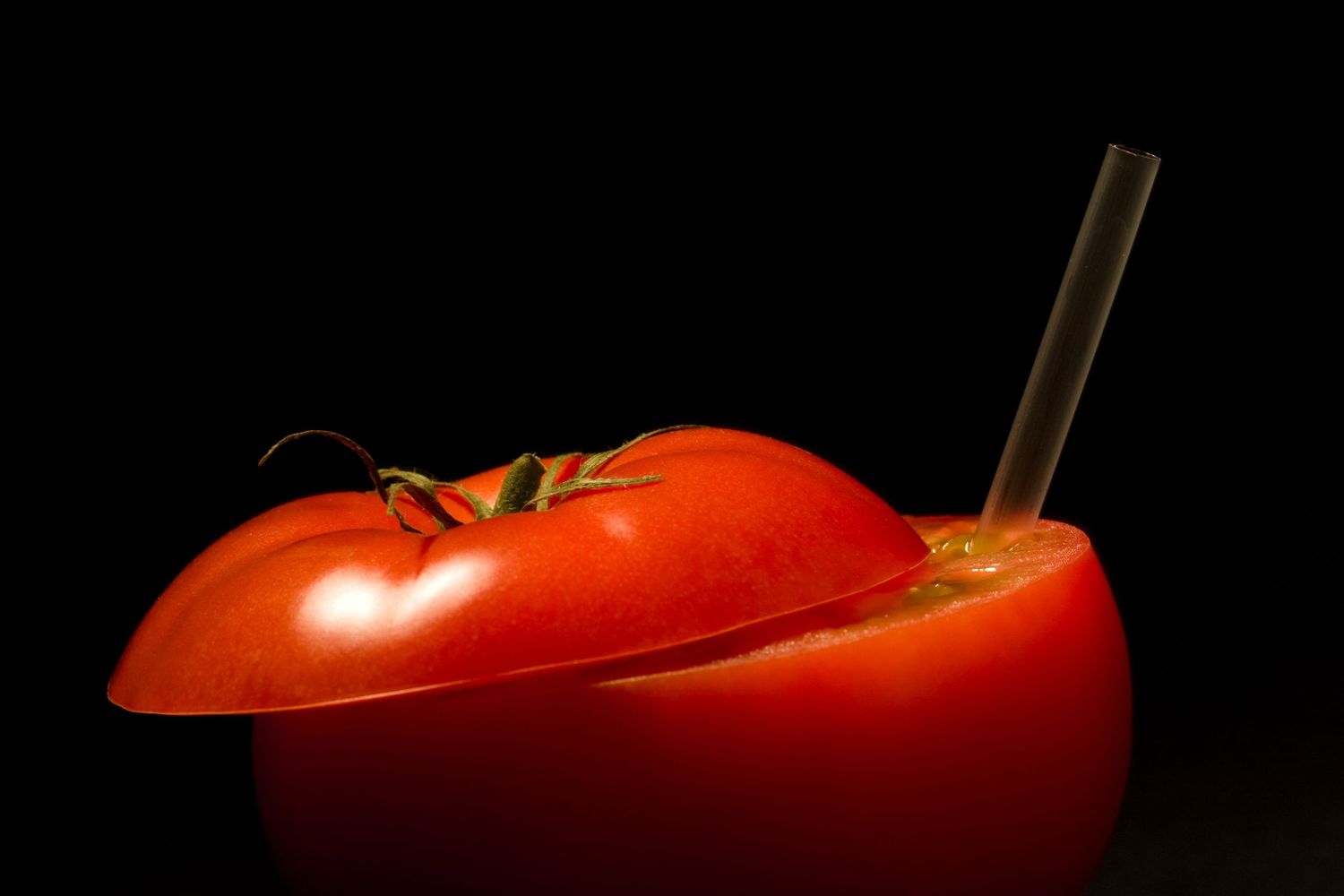 Conceptual fine art of a sliced tomato with a straw, creating the illusion of drinking fresh tomato juice on a black background.