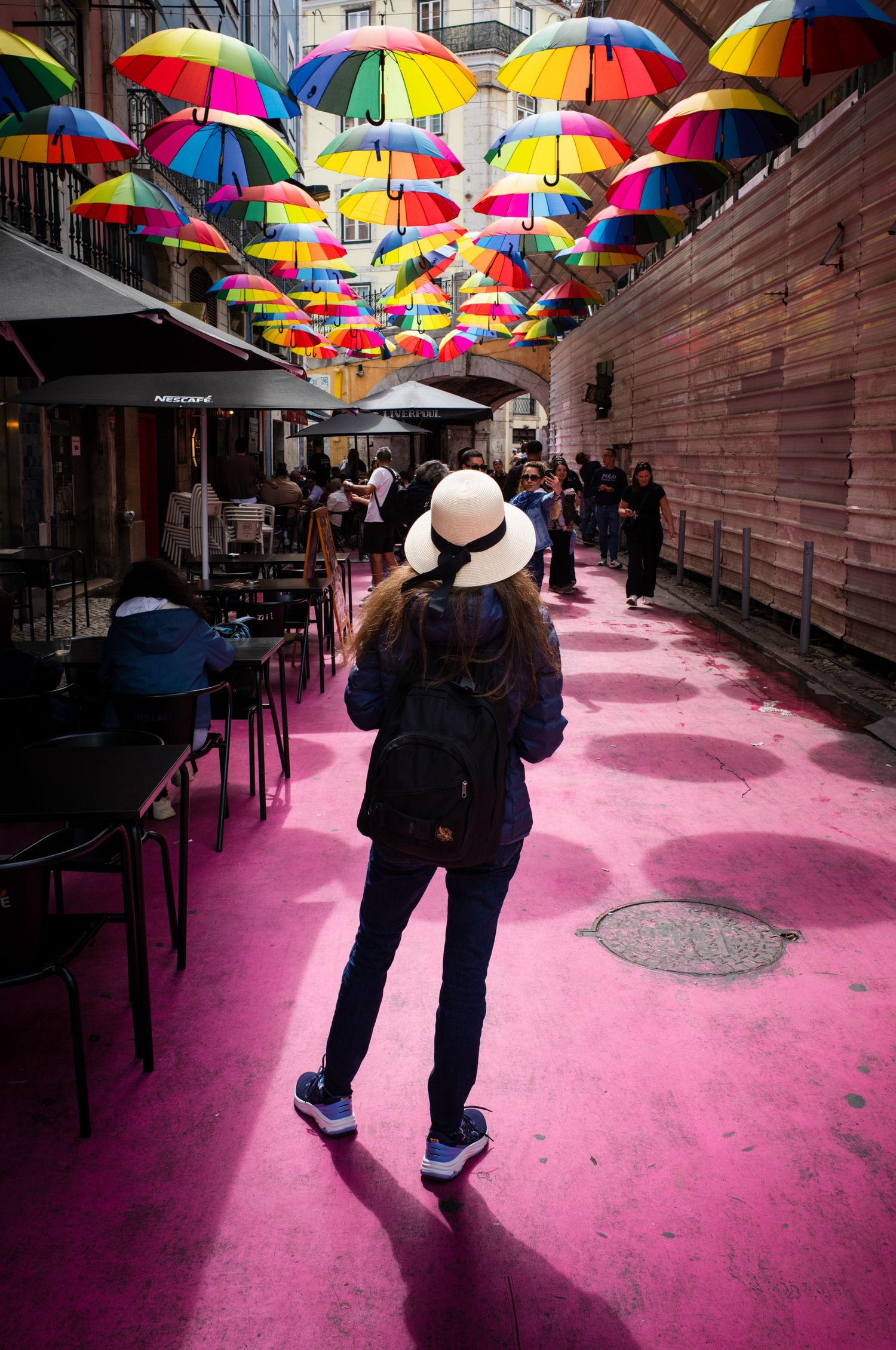 Woman in a white sun hat walks beneath colorful umbrellas on Lisbon's Pink Street, blending vibrant travel and street photography.
