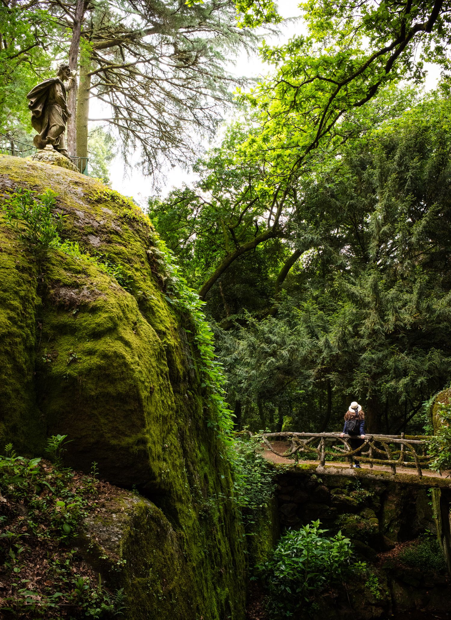 Woman in a white hat crossing a rustic bridge in Bom Jesus do Monte gardens, Braga, Portugal, surrounded by lush forest.