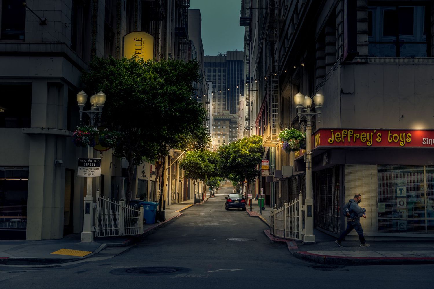 Blue hour view of a quiet downtown San Francisco alley framed by trees, glowing streetlights and a lone pedestrian.