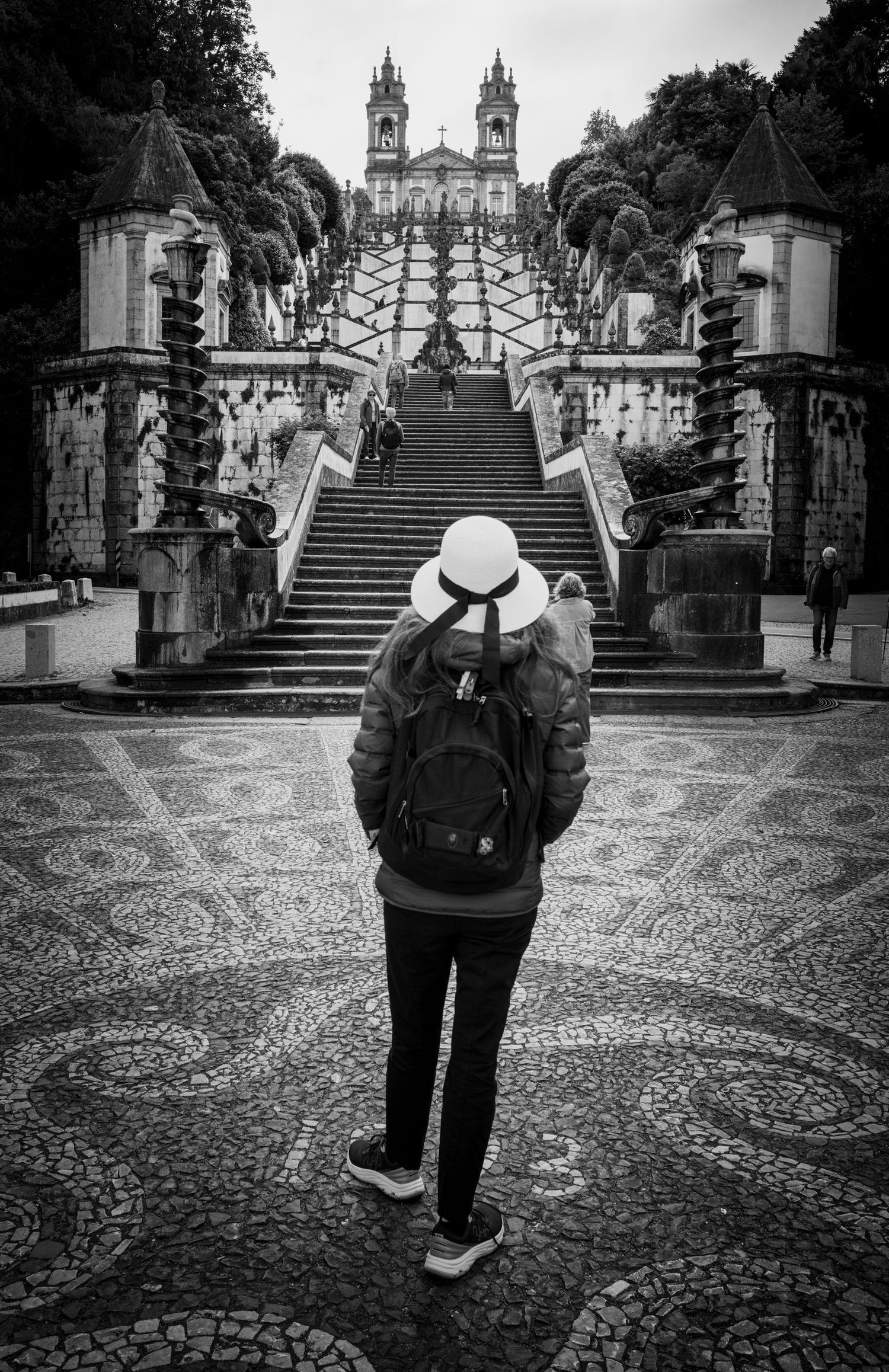 Woman in white hat faces the iconic Bom Jesus do Monte staircase in Braga, Portugal, captured in dramatic black and white.