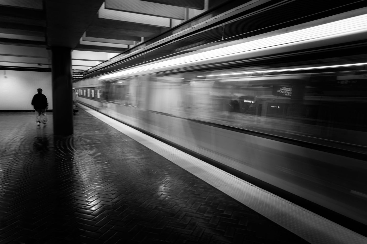 Long-exposure black-and-white photo of a moving San Francisco BART train passing a lone commuter on the platform.