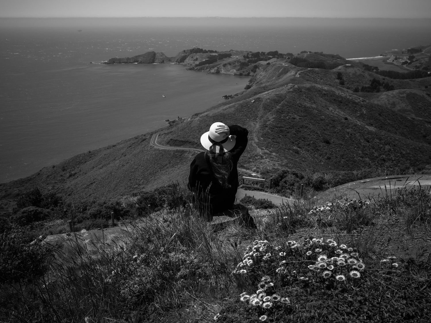 Woman in a white hat overlooks the Marin Headlands and Pacific coastline near windy San Francisco in a black-and-white landscape.