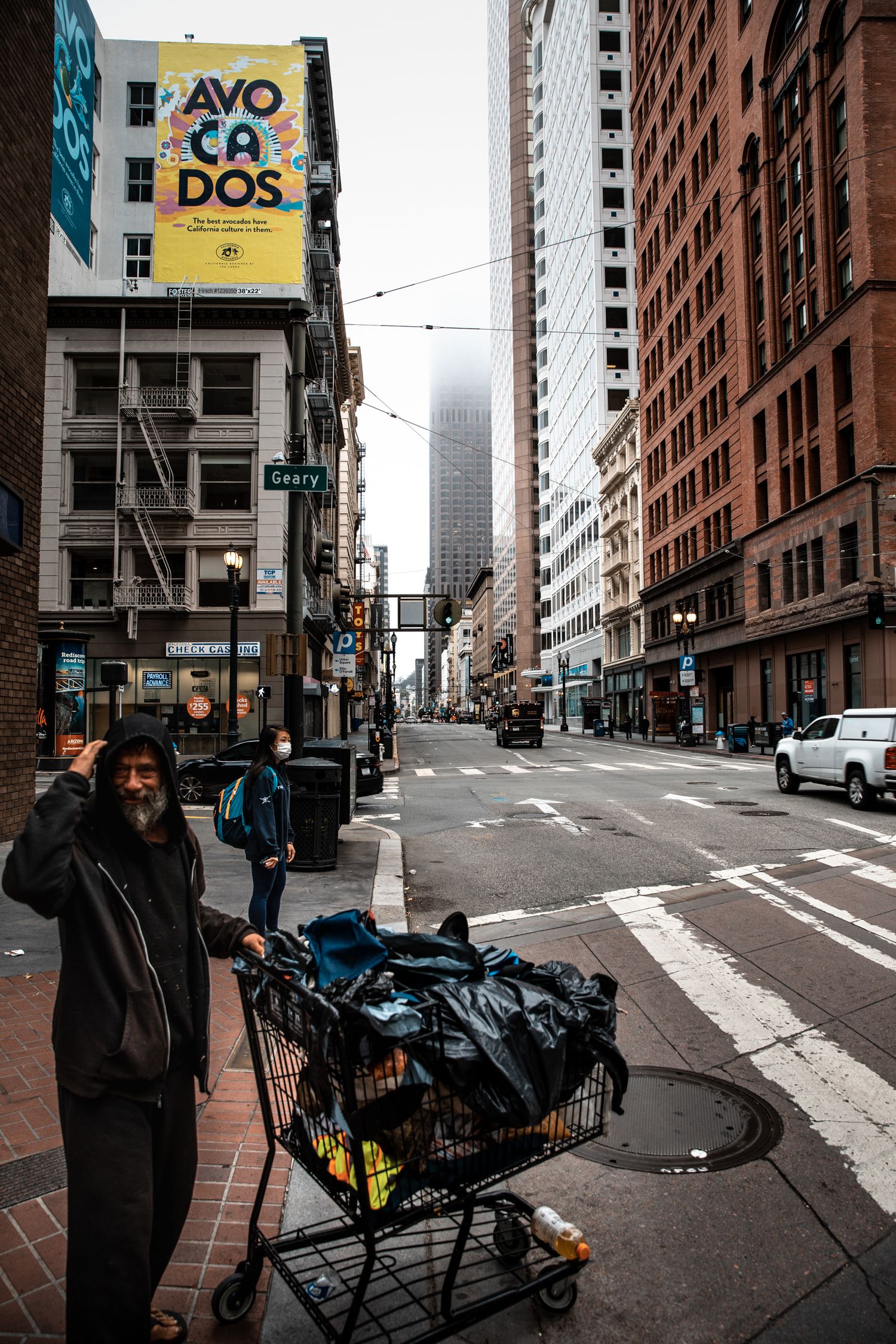 Man with shopping cart on Geary Street beneath foggy Financial District towers in a candid San Francisco street scene.