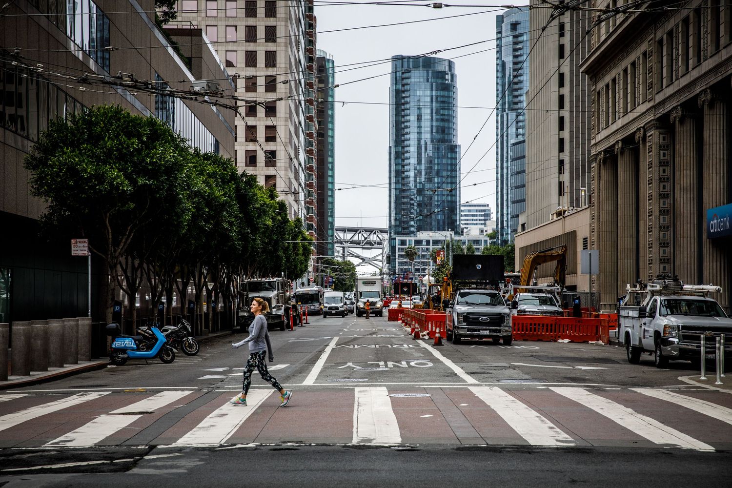 Pedestrian crossing a downtown San Francisco intersection framed by skyscrapers, streetcar wires, and urban city life.