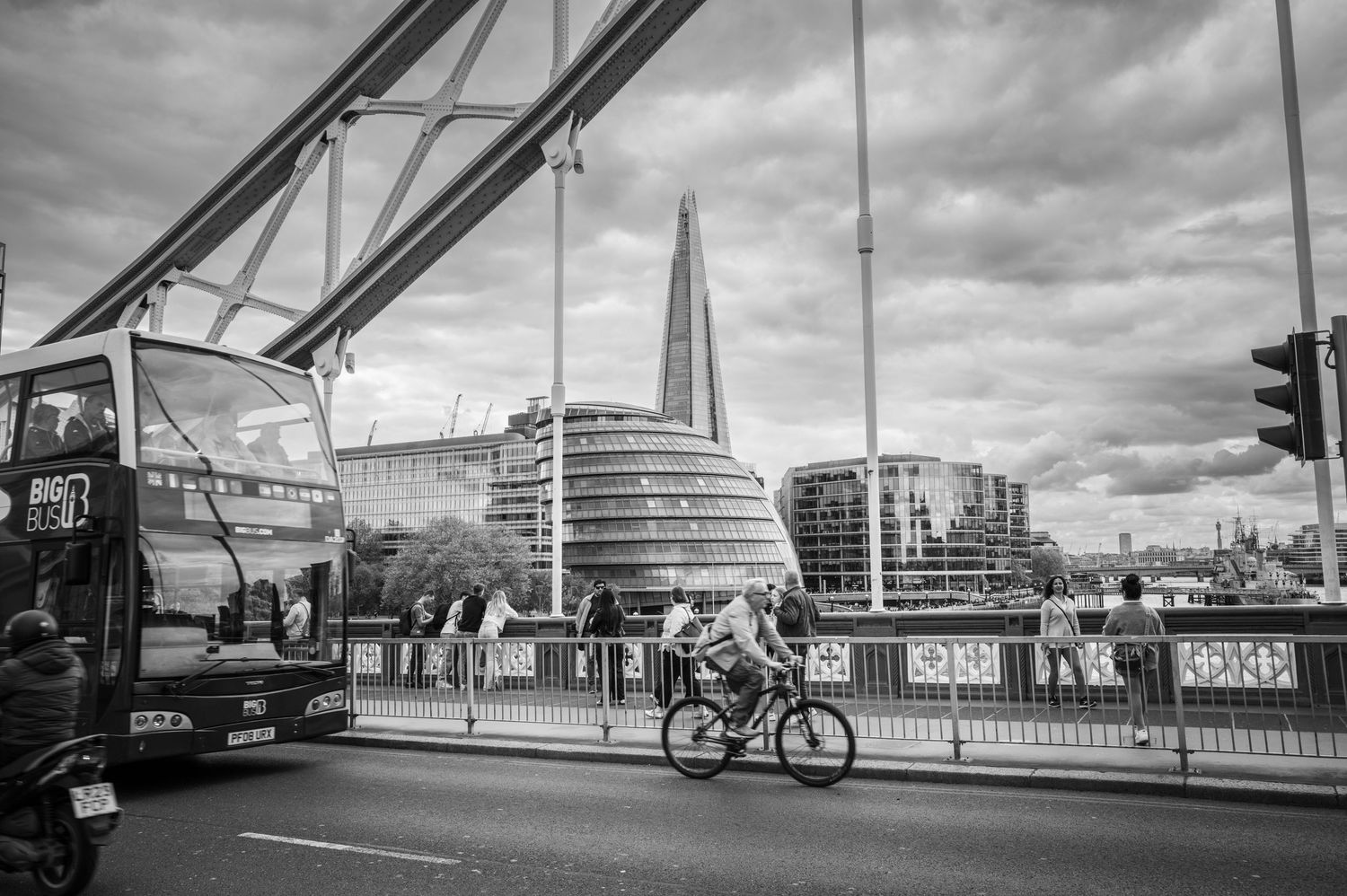 Black-and-white street scene on Tower Bridge in London with a cyclist, double-decker bus, The Shard, and dramatic storm clouds.
