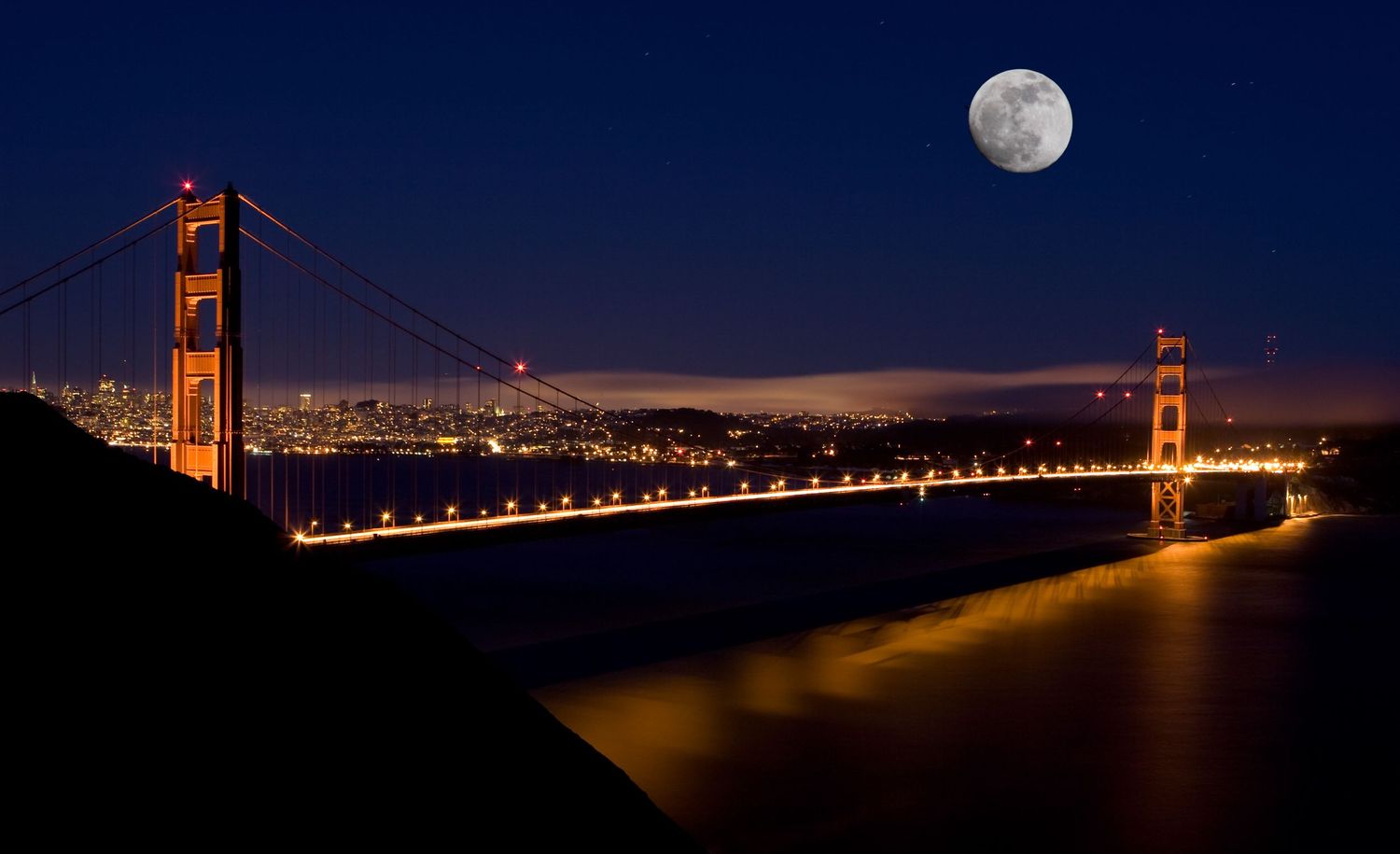 Golden Gate Bridge illuminated at night beneath a full moon with San Francisco skyline glowing across the bay in a long exposure.