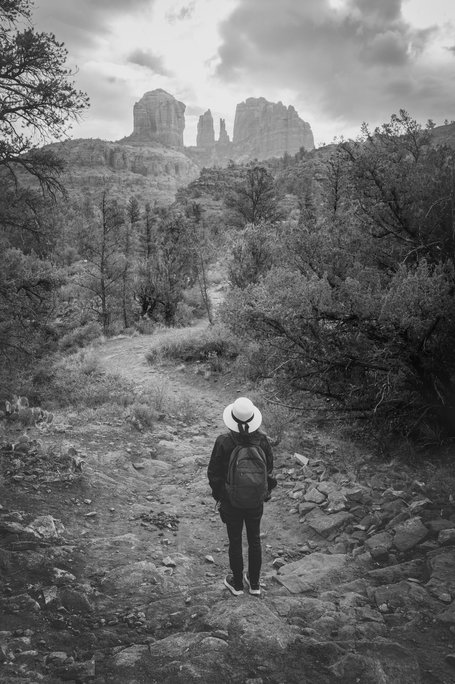 Traveler in a white hat overlooks Cathedral Rock along a Sedona trail beneath dramatic storm clouds in black and white.