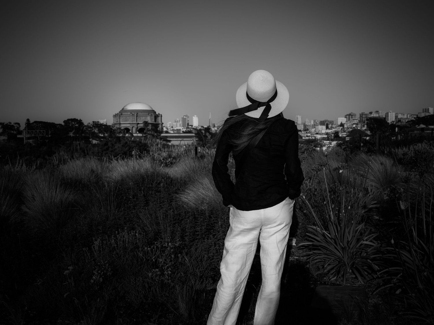 Woman in a sun hat overlooks the Palace of Fine Arts from Chrissy Field, San Francisco, in a contemplative black-and-white scene.