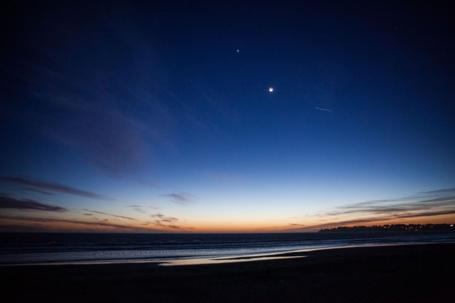 Crescent moon and bright planets shine over the Santa Cruz, California coastline at twilight above the peaceful Pacific Ocean.