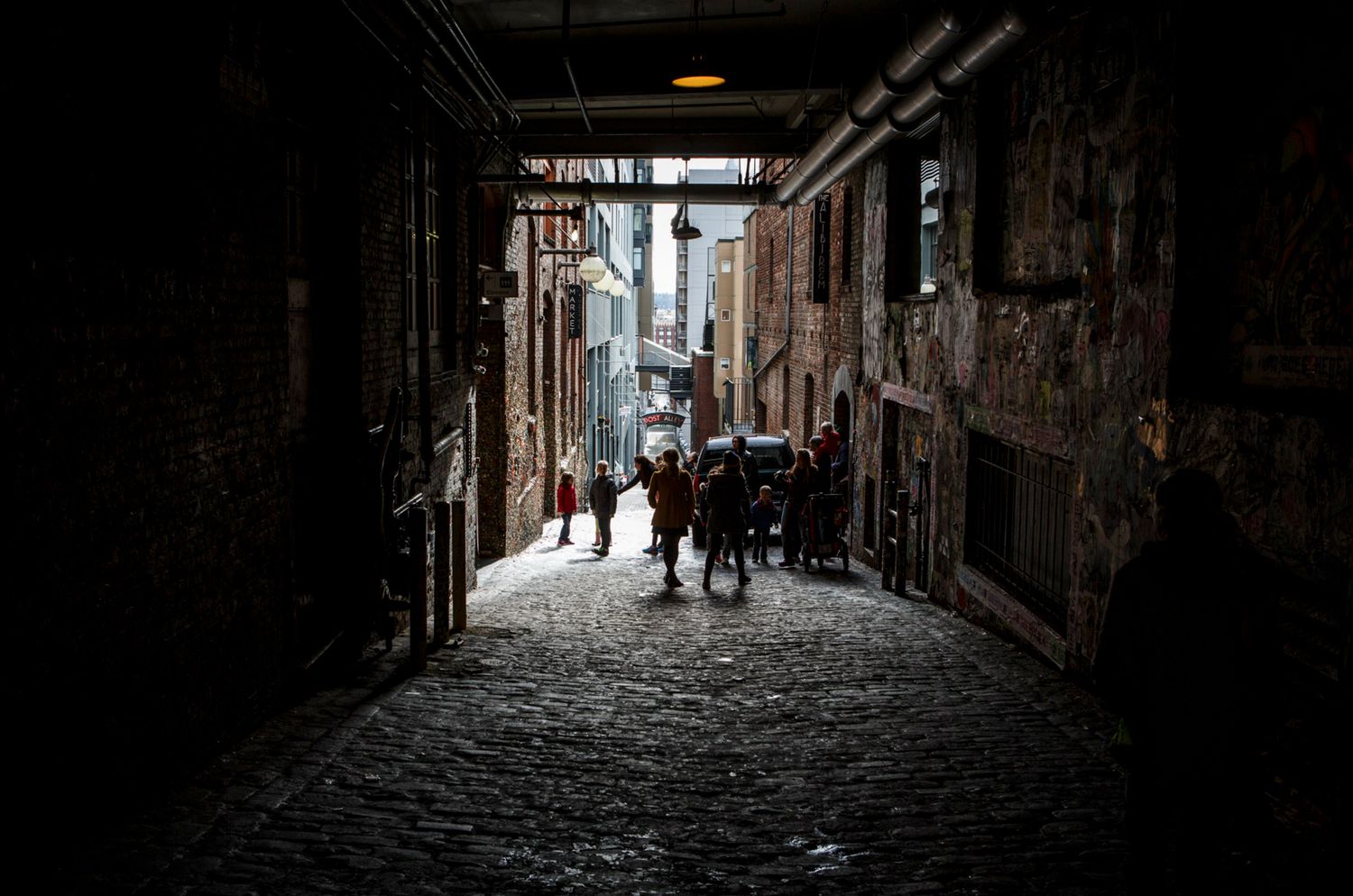 Silhouetted visitors walk through Gum Wall Alley at Seattle's Pike Place Market, framed by dramatic light and shadow.